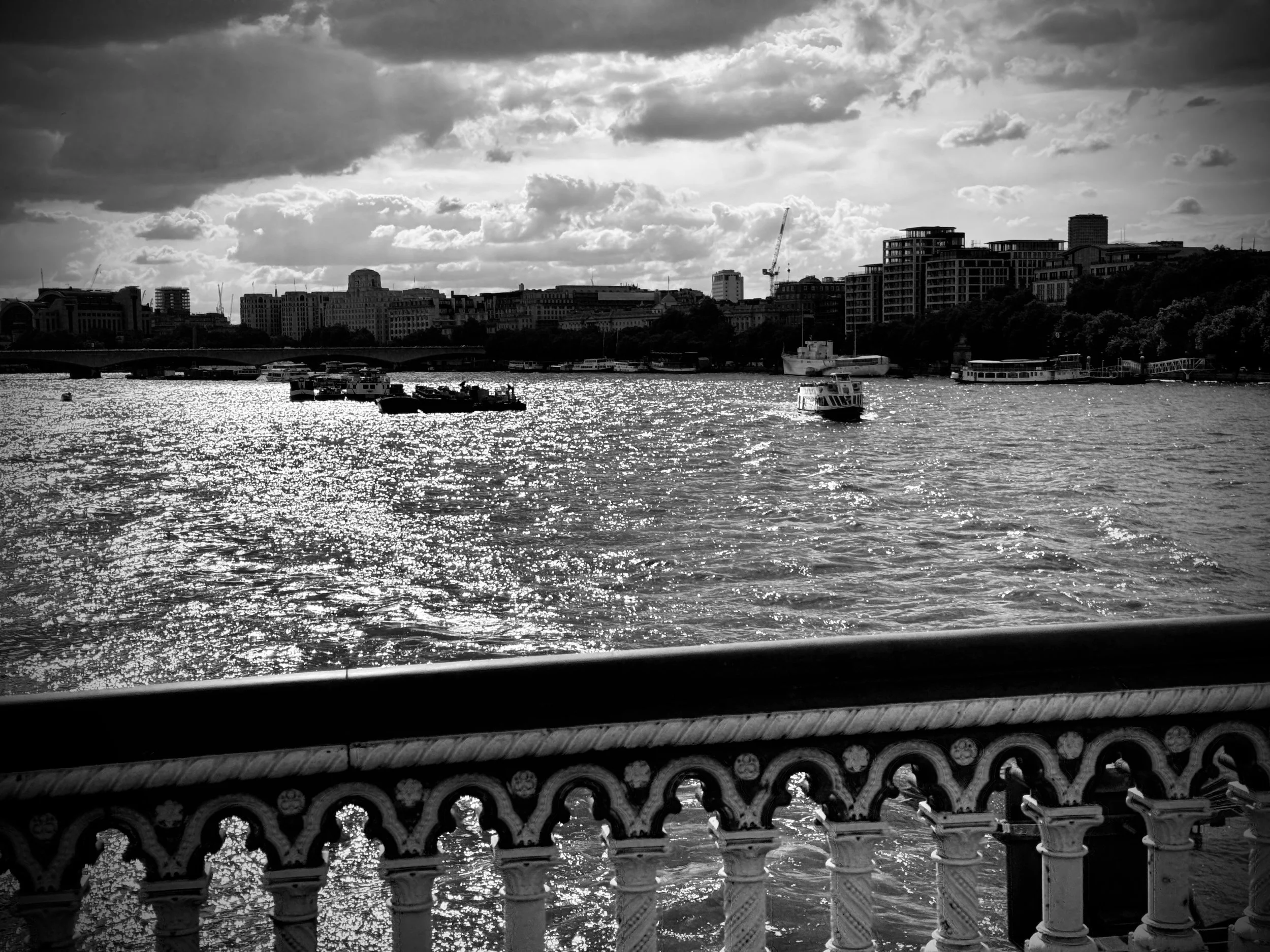 Monochrome cityscape showing cranes and boats beneath a brooding sky — an industrial skyline viewed from an ornate riverside balcony.