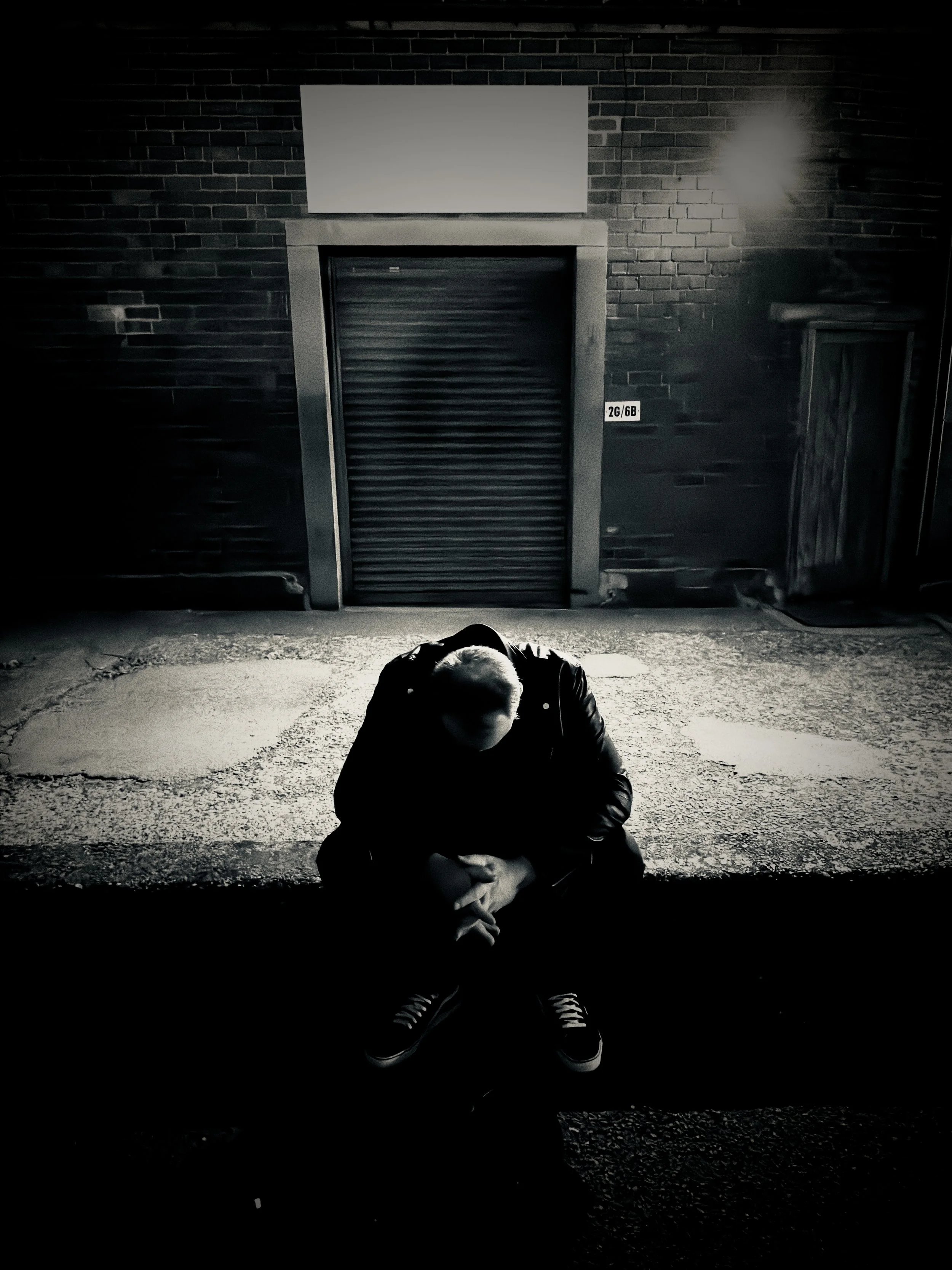 Monochrome night‑time portrait of UK musician Paul Arden sitting on a curb outside a brick building, head bowed in reflection. The single wall light casts dramatic shadows, creating a moody, urban atmosphere that captures his contemplative visual sty