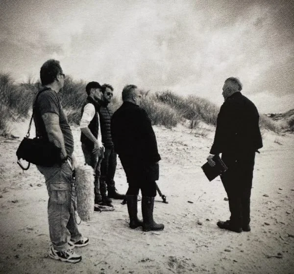 Film crew gathered on a windswept beach, listening to direction from the director holding notes — a quiet moment of collaboration before shooting.