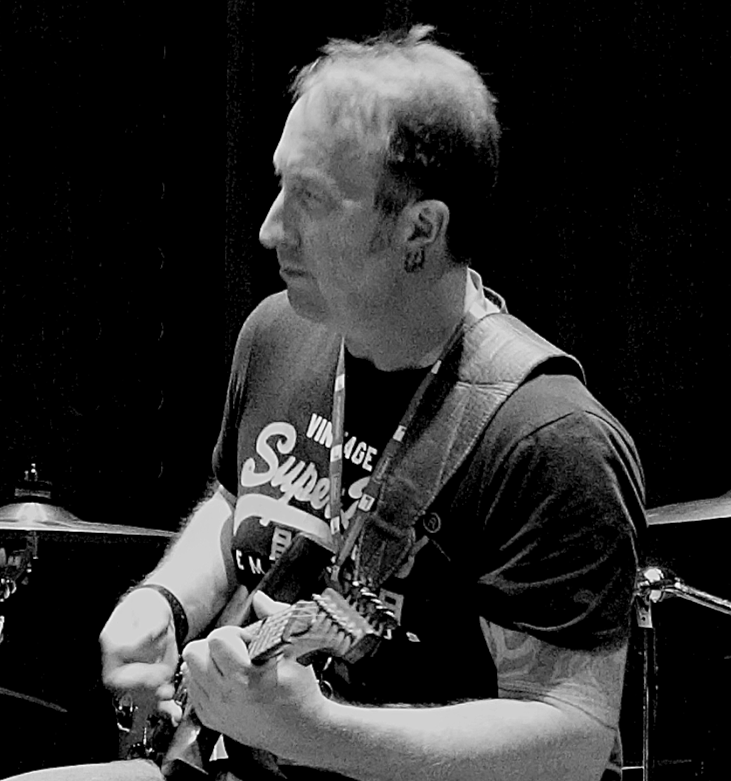 A person with short, light-colored hair playing an electric guitar, wearing a dark T-shirt and a lanyard around their neck, while sitting in front of a drum set.