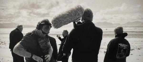 Black‑and‑white shot of a sound team preparing gear on a coastal set, adjusting microphones and boom poles against the backdrop of open sea.