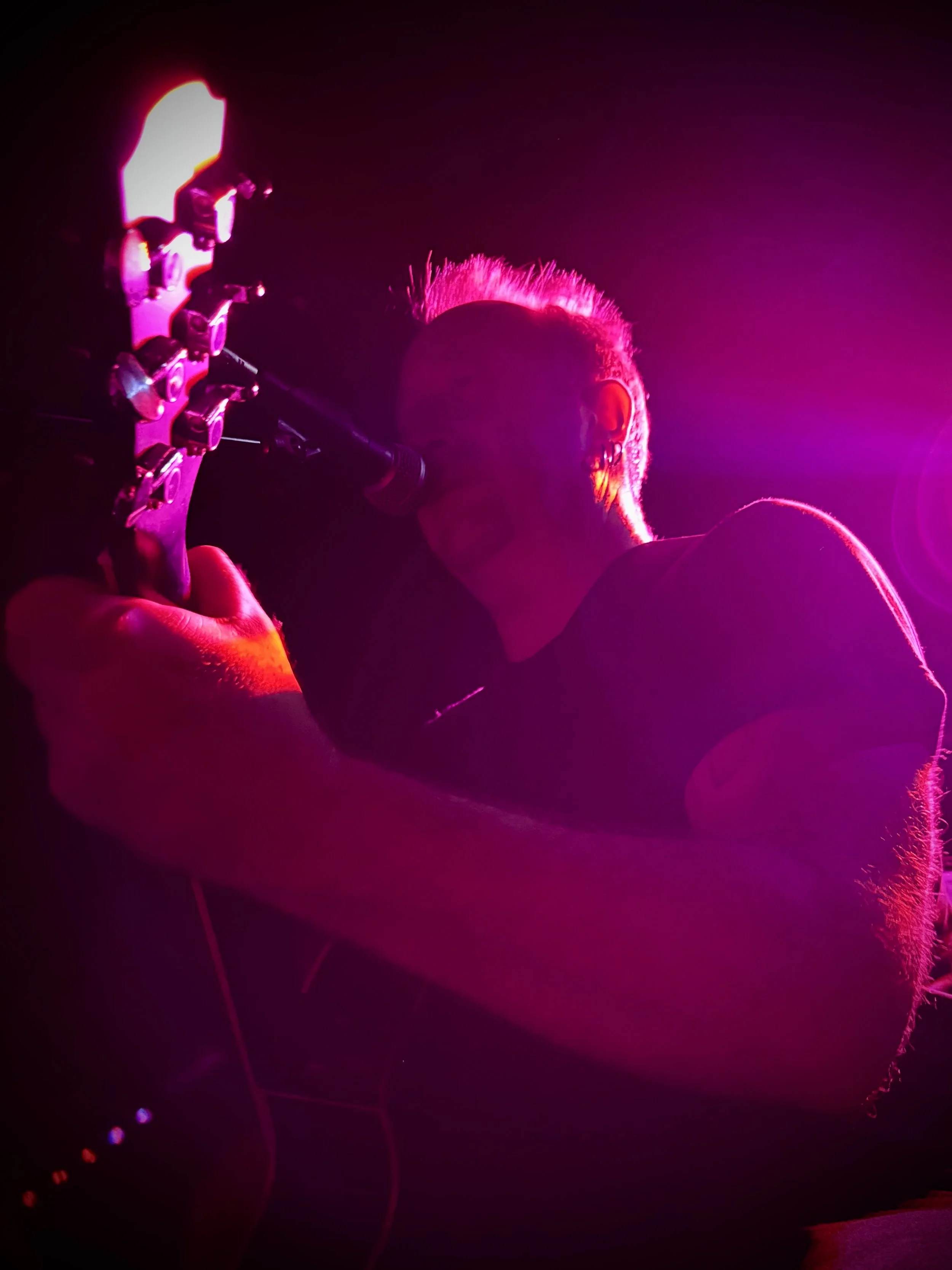 Monochrome‑style live performance photograph of UK musician Paul Arden playing guitar under vivid purple and pink stage lighting. The shot captures the intensity and atmosphere of a live show, highlighting his expressive performance style and cinemat
