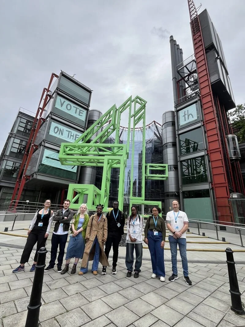 Seven people gathered before the Millennium Monument in London, framed by modern architecture and a bold campaign message — a moment of civic energy and collaboration.