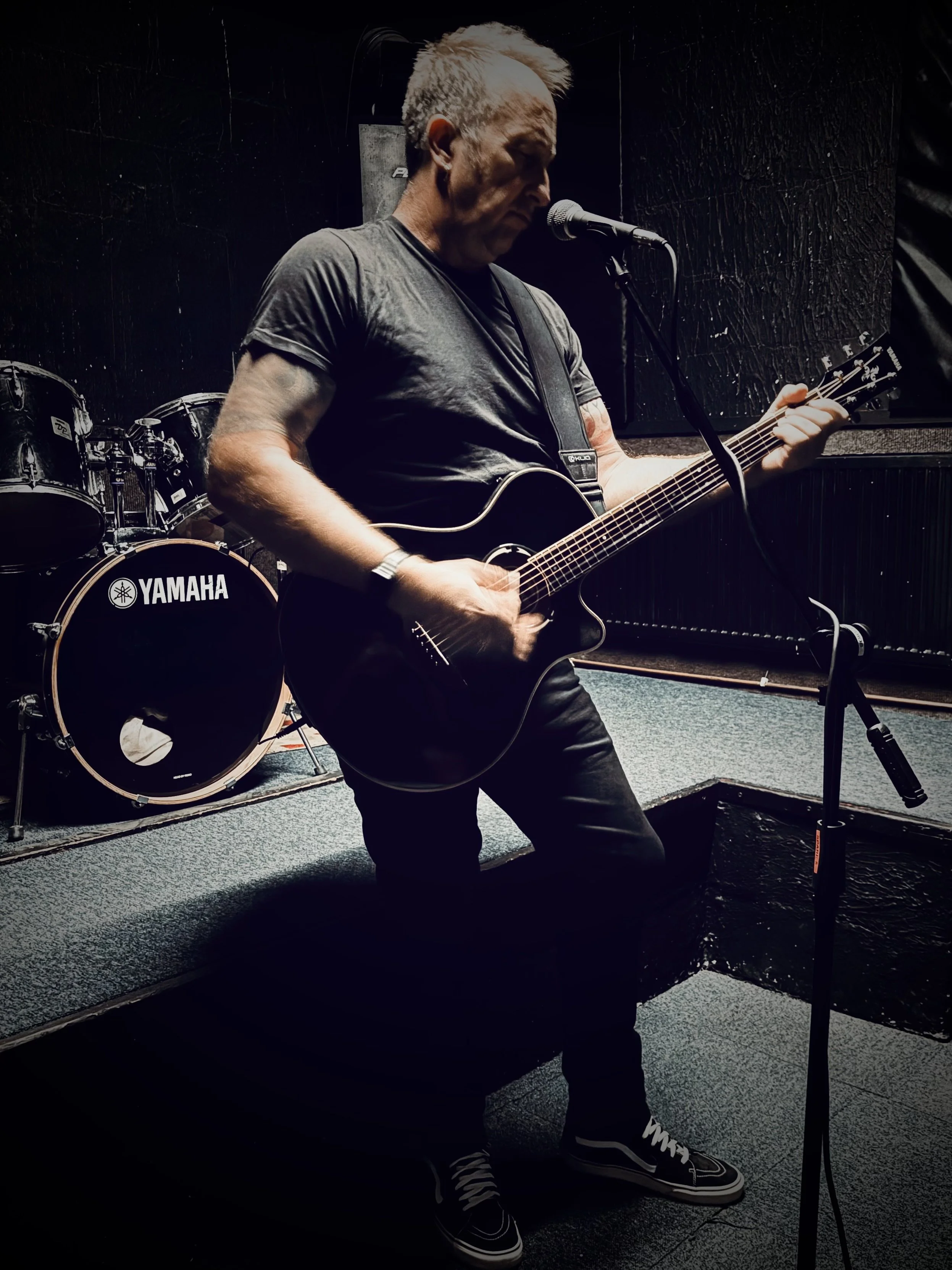Studio rehearsal photograph of UK musician Paul Arden performing live vocals and acoustic guitar in a dark, soundproofed room. A Yamaha drum kit sits behind him, capturing the raw, cinematic atmosphere of his creative process and rehearsal sessions.