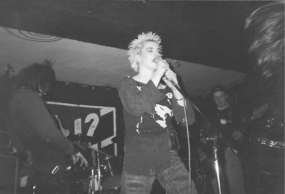 Dynamic live performance featuring a blonde vocalist at centre stage, backed by guitar and drums — raw energy captured in monochrome.