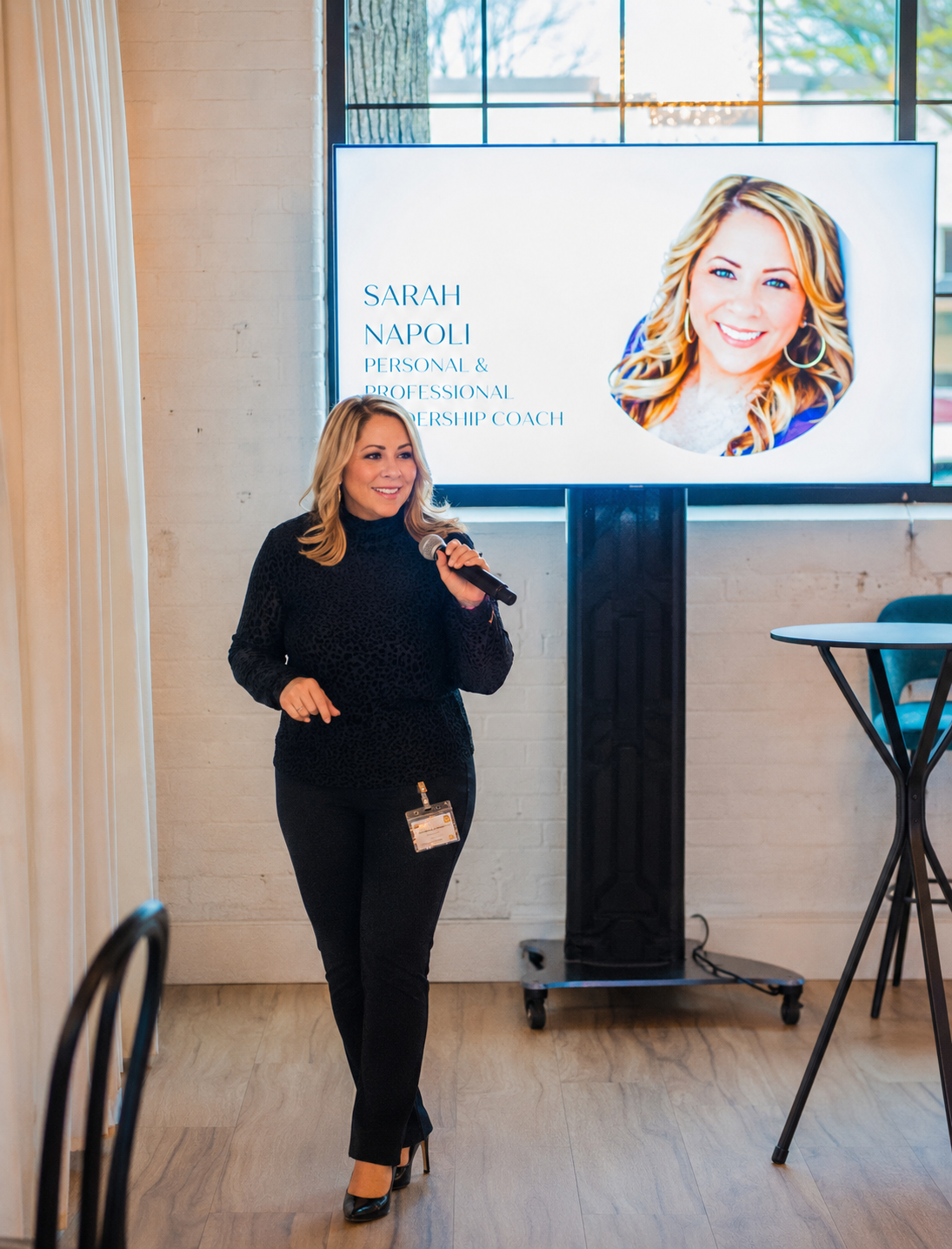 A woman giving a presentation appears to be at a conference or seminar. She is holding a microphone, has blonde hair, and is dressed in black. Behind her, a screen displays her name, Sarah Napoli, and her titles as a personal and professional leadership coach. The screen also shows a large portrait of her smiling.