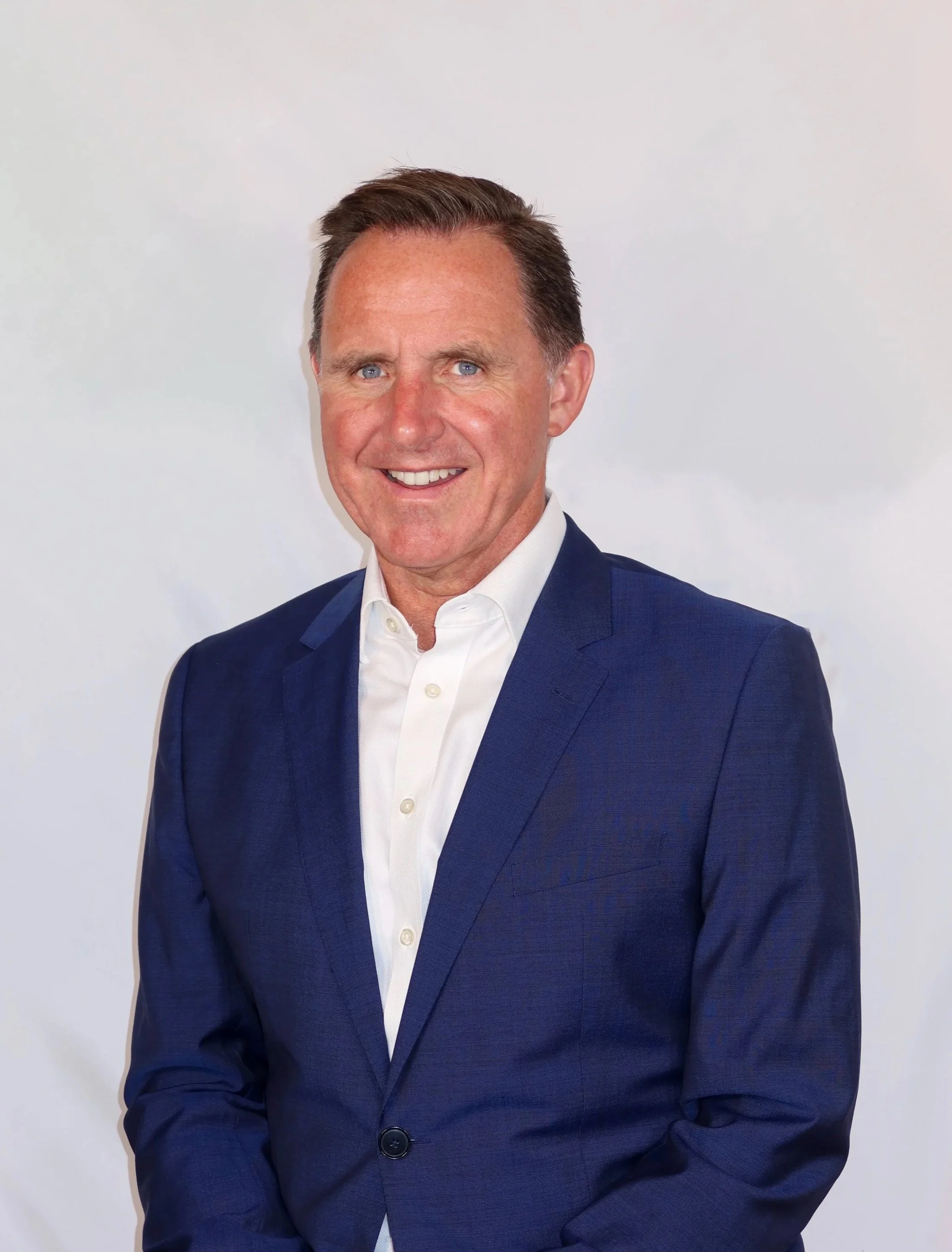 Man with short brown hair, wearing a dark blue suit jacket and a white dress shirt, smiling against a plain white background.