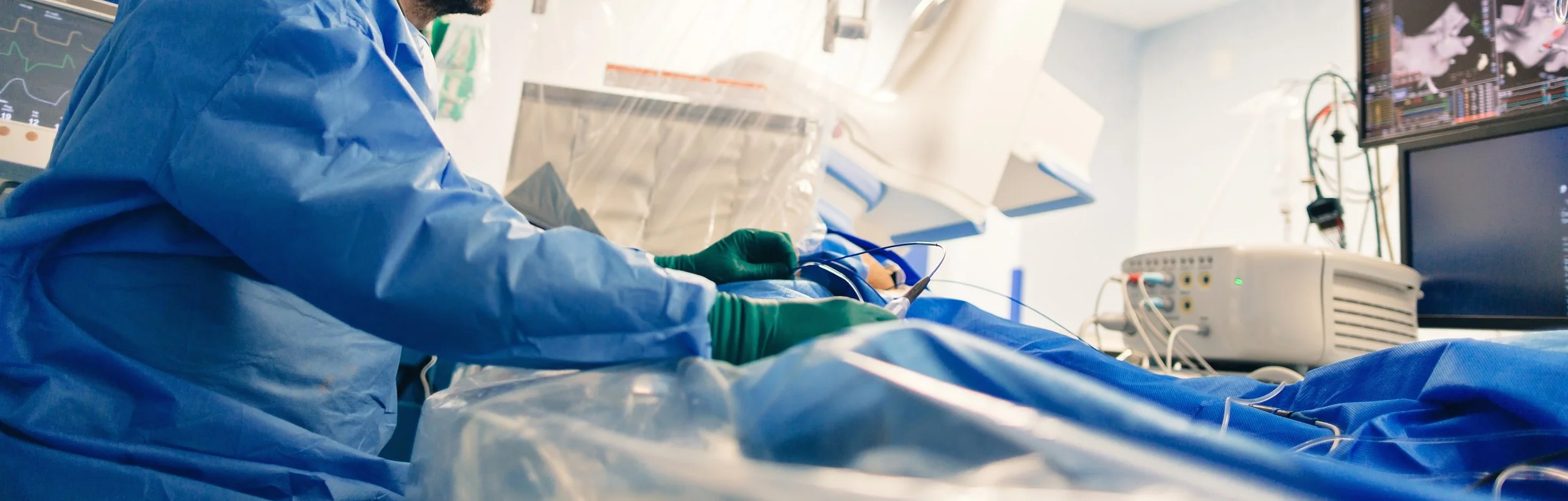 Medical professional in blue protective gown and green gloves performs a procedure on a patient in a hospital setting, with monitors displaying vital signs in the background.