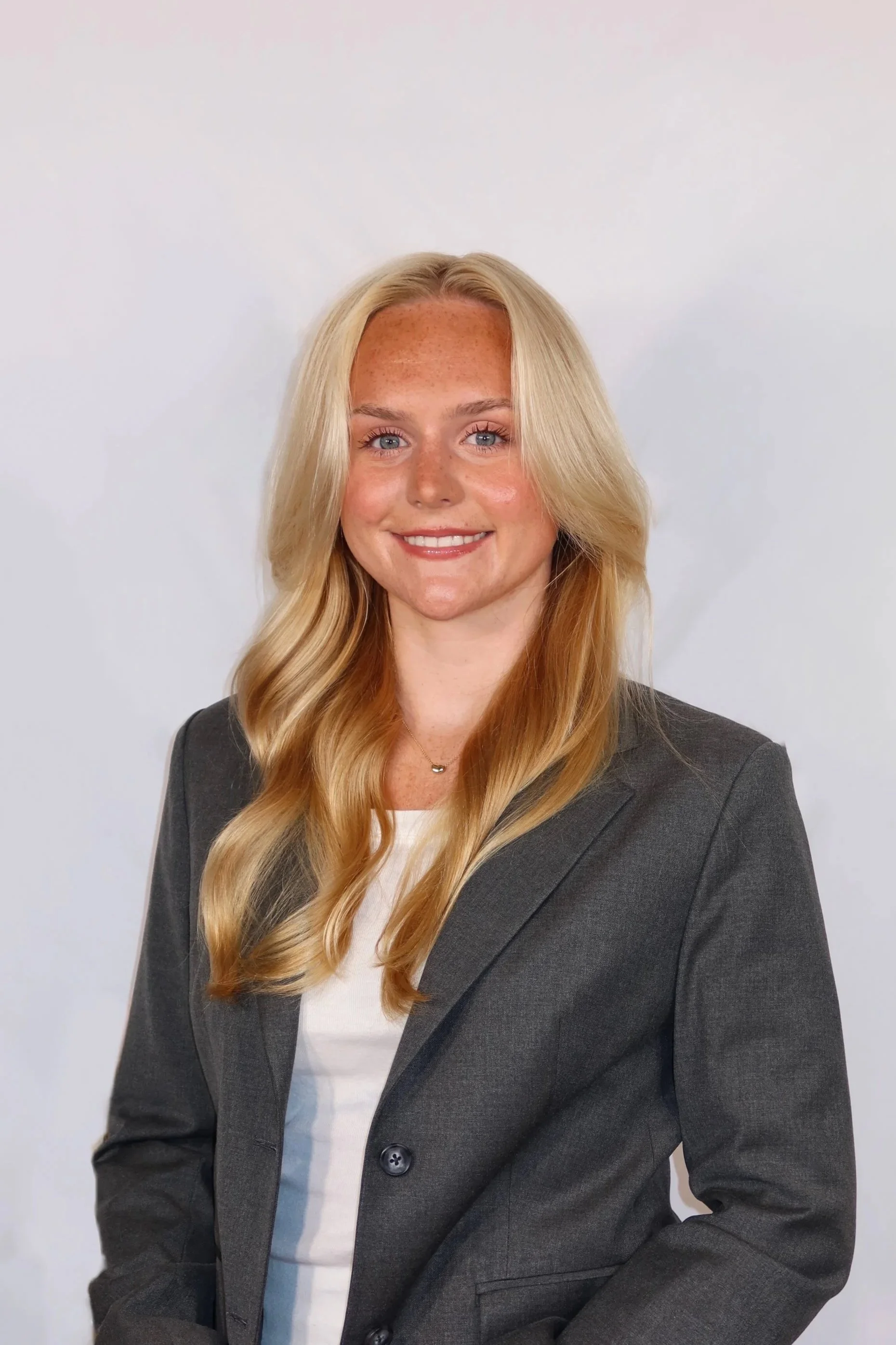 A woman with long, wavy blonde hair and light skin, smiling, wearing a dark gray blazer and a white top, standing against a plain white background.