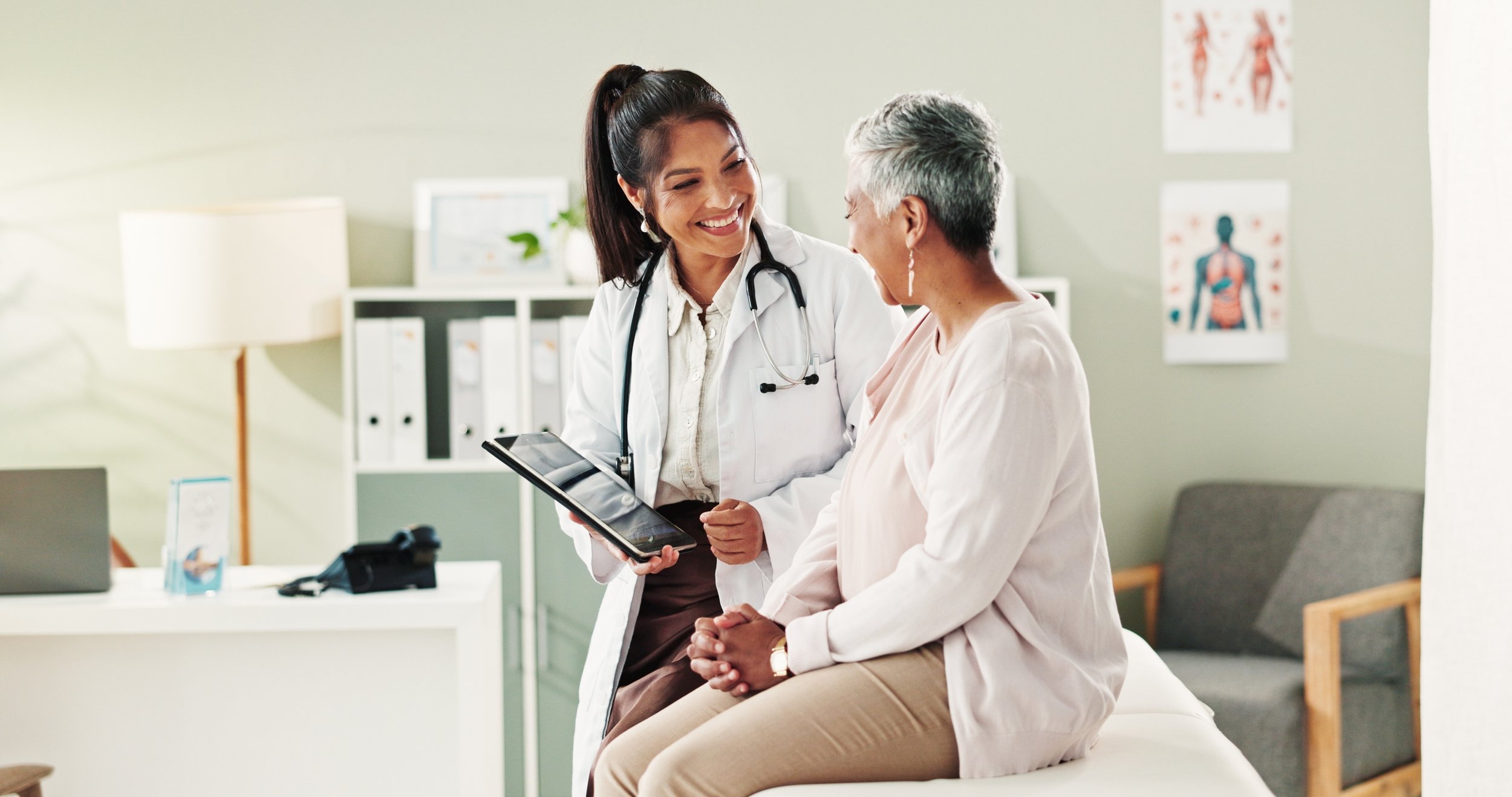 Female doctor smiling and talking to an elderly female patient in a medical office, holding a tablet.