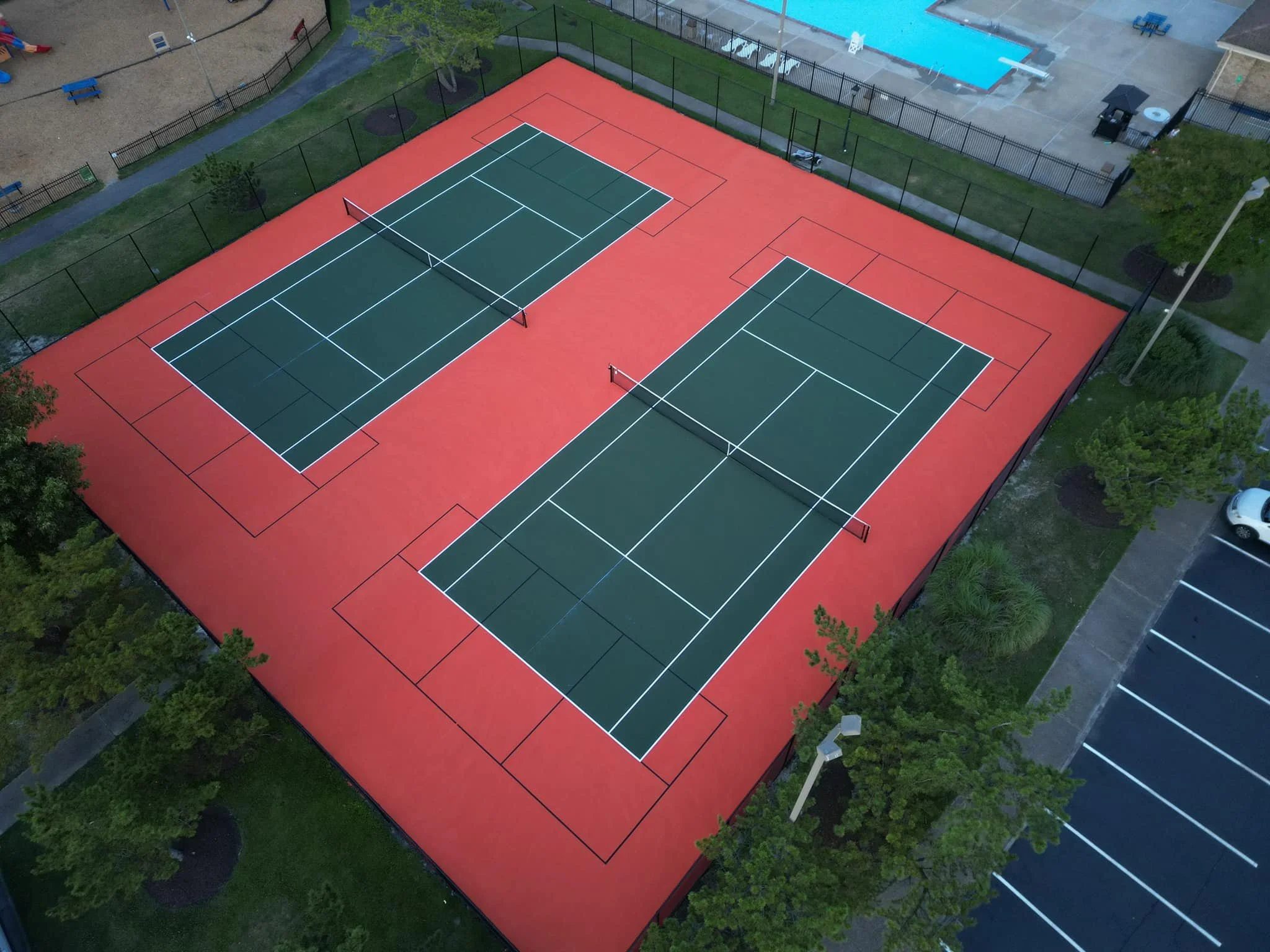 Aerial view of a tennis court with green playing surface and white lines, surrounded by a red playing area, enclosed by a black fence. There are two tennis nets in the center, and a school or community area with a swimming pool and a playground nearby.