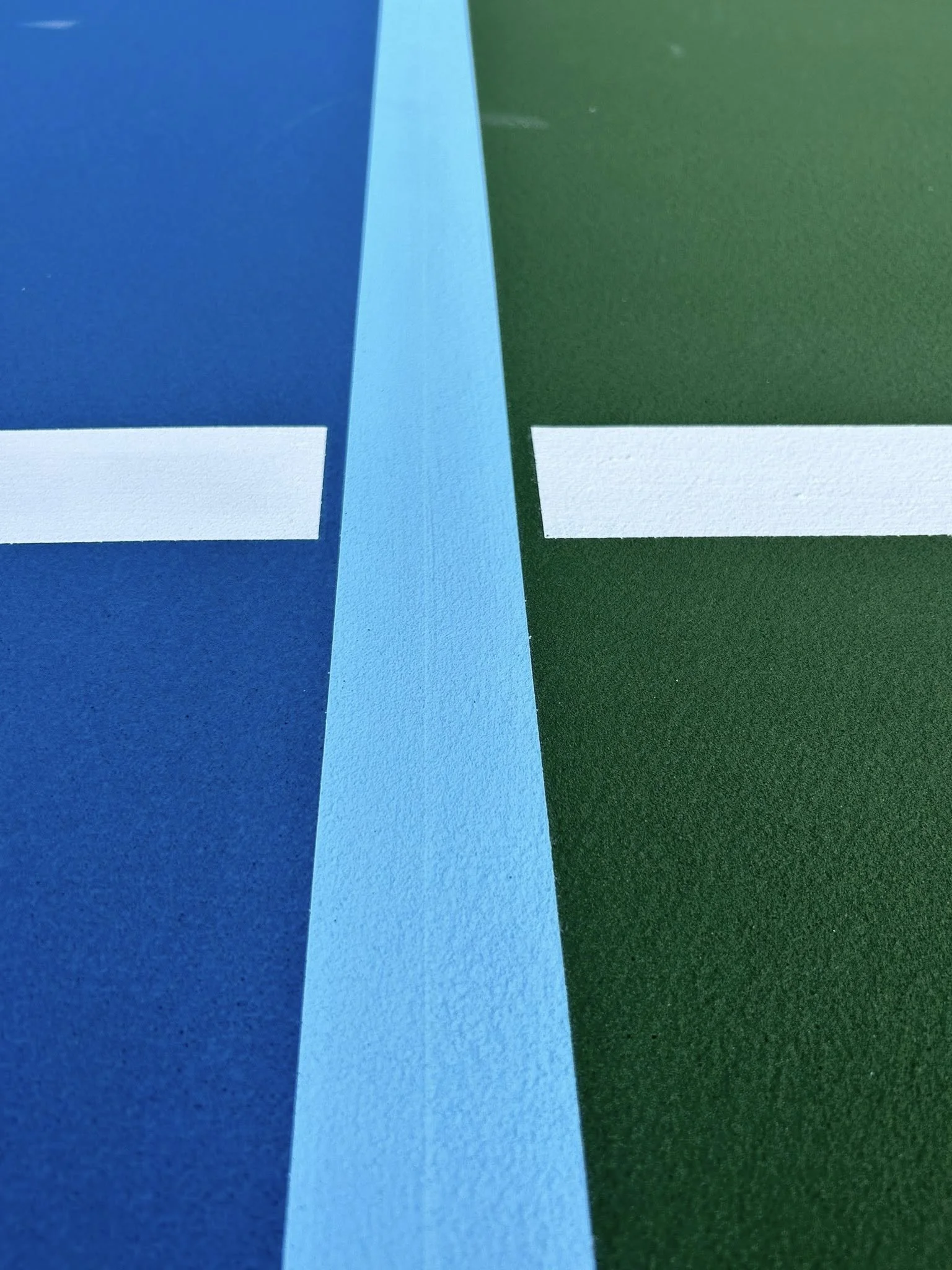 Close-up of a tennis court surface with painted lines, showing blue on the left side and green on the right side.