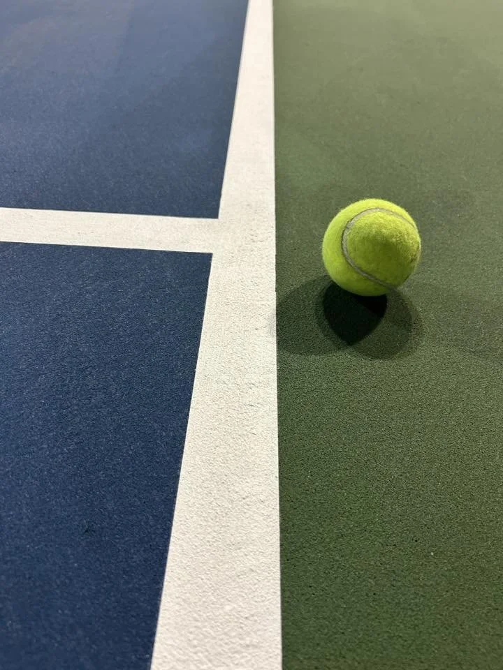 A yellow tennis ball resting on a green tennis court surface near a white boundary line, with a blue court visible adjacent to the green court.