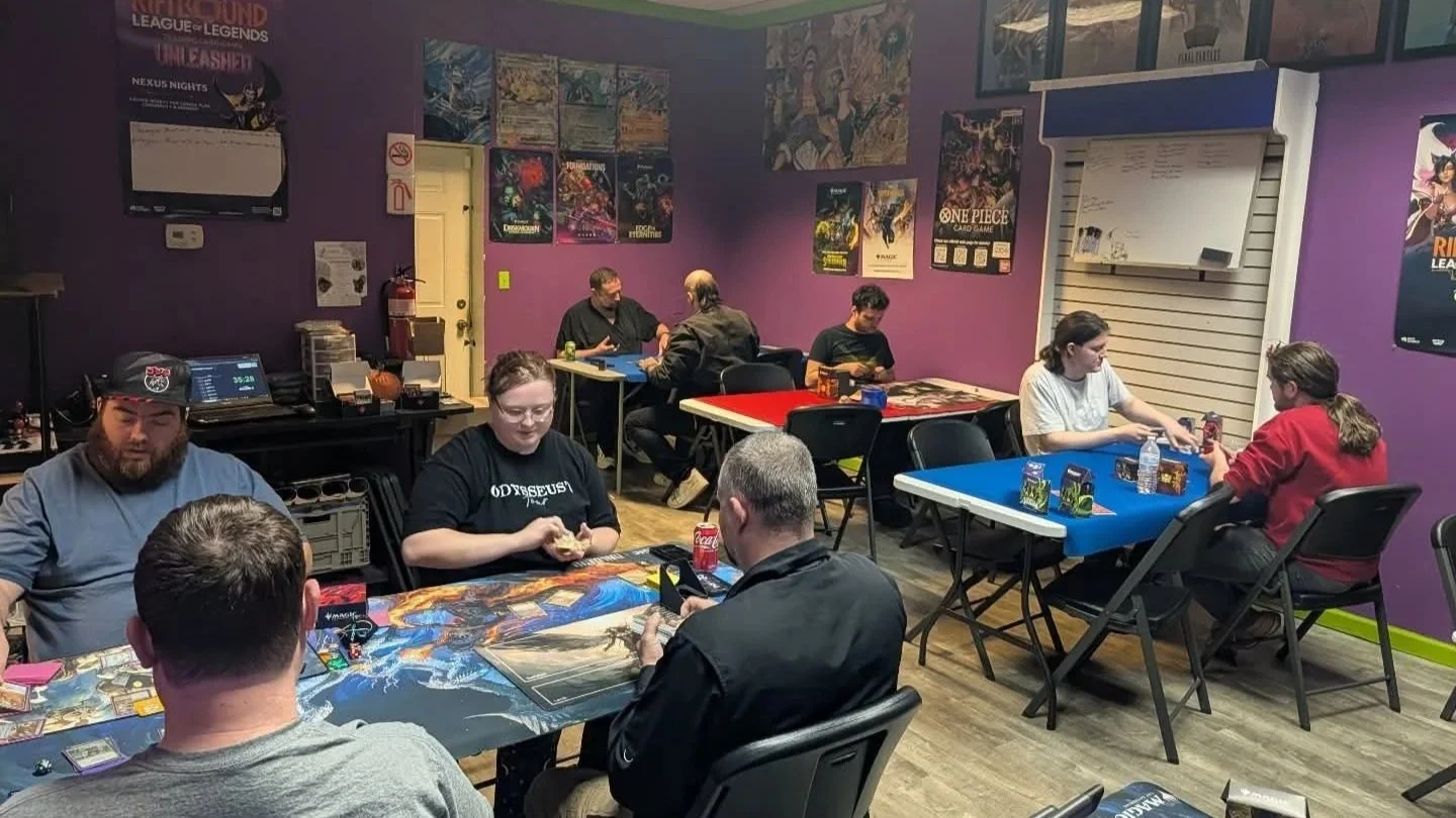 Players gather around tables at a community game store, playing tabletop card and board games in a purple-walled room decorated with gaming posters.
