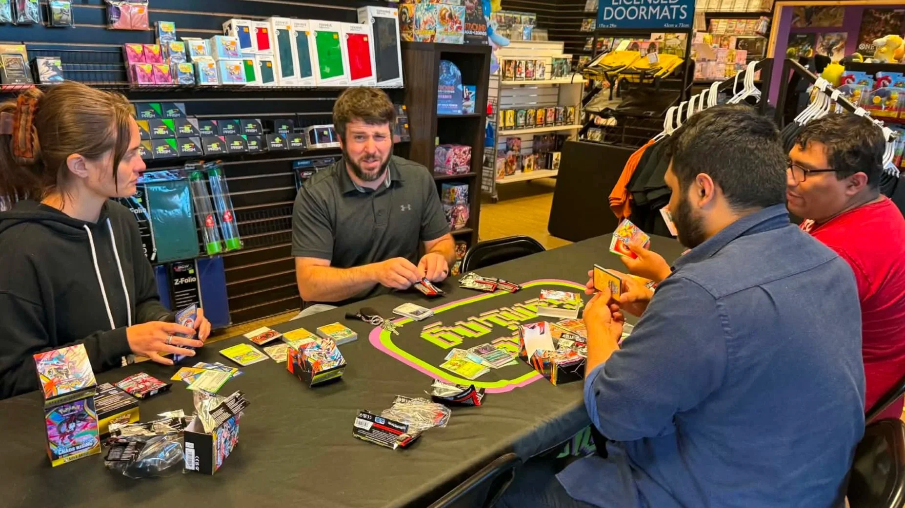 Four people play a Pokémon trading card game at a tabletop in a hobby store surrounded by cards and collectibles.