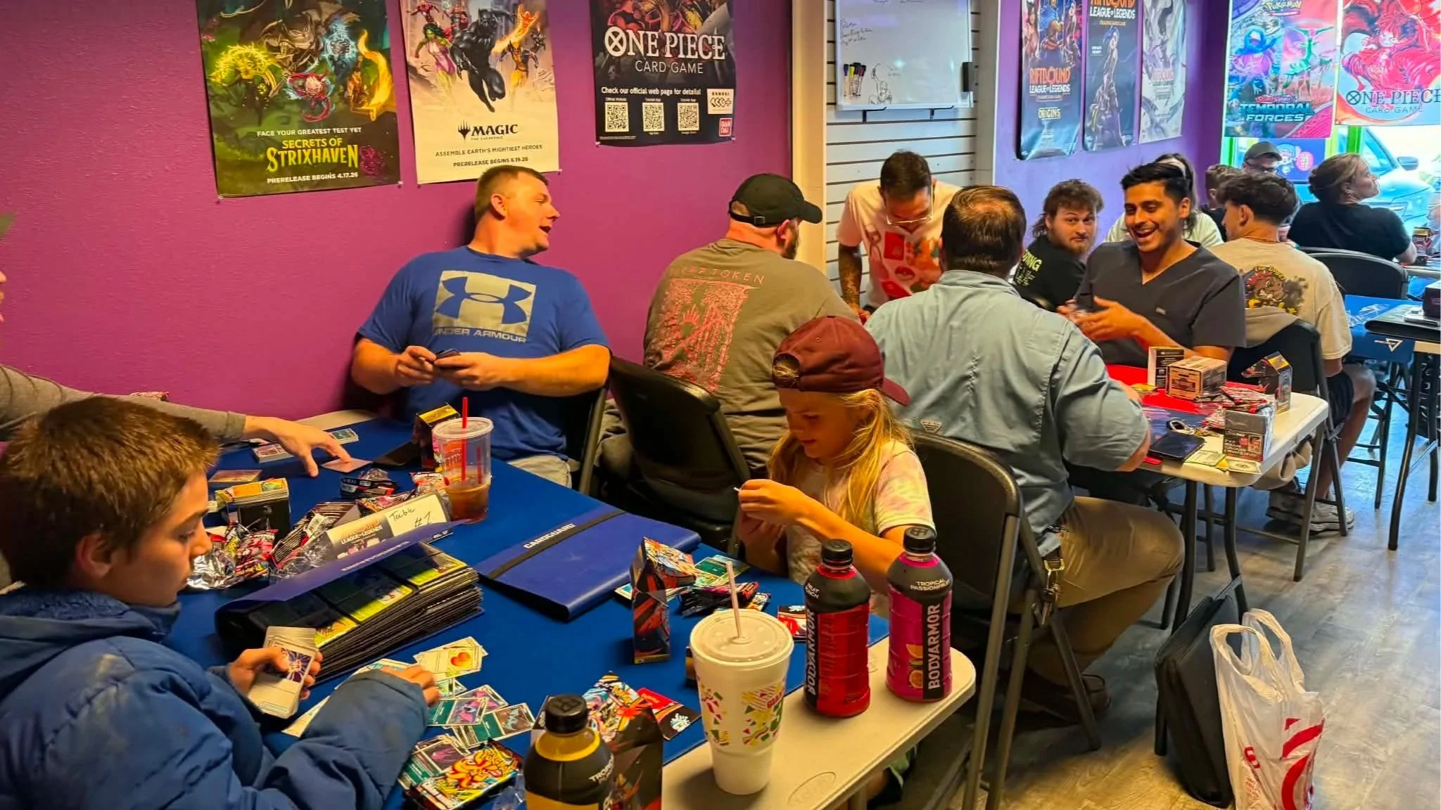 Players gather at tables for a trading card game tournament in a colorful card shop decorated with Magic: The Gathering, Pokémon, Yu-Gi-Oh!, and One Piece posters.
