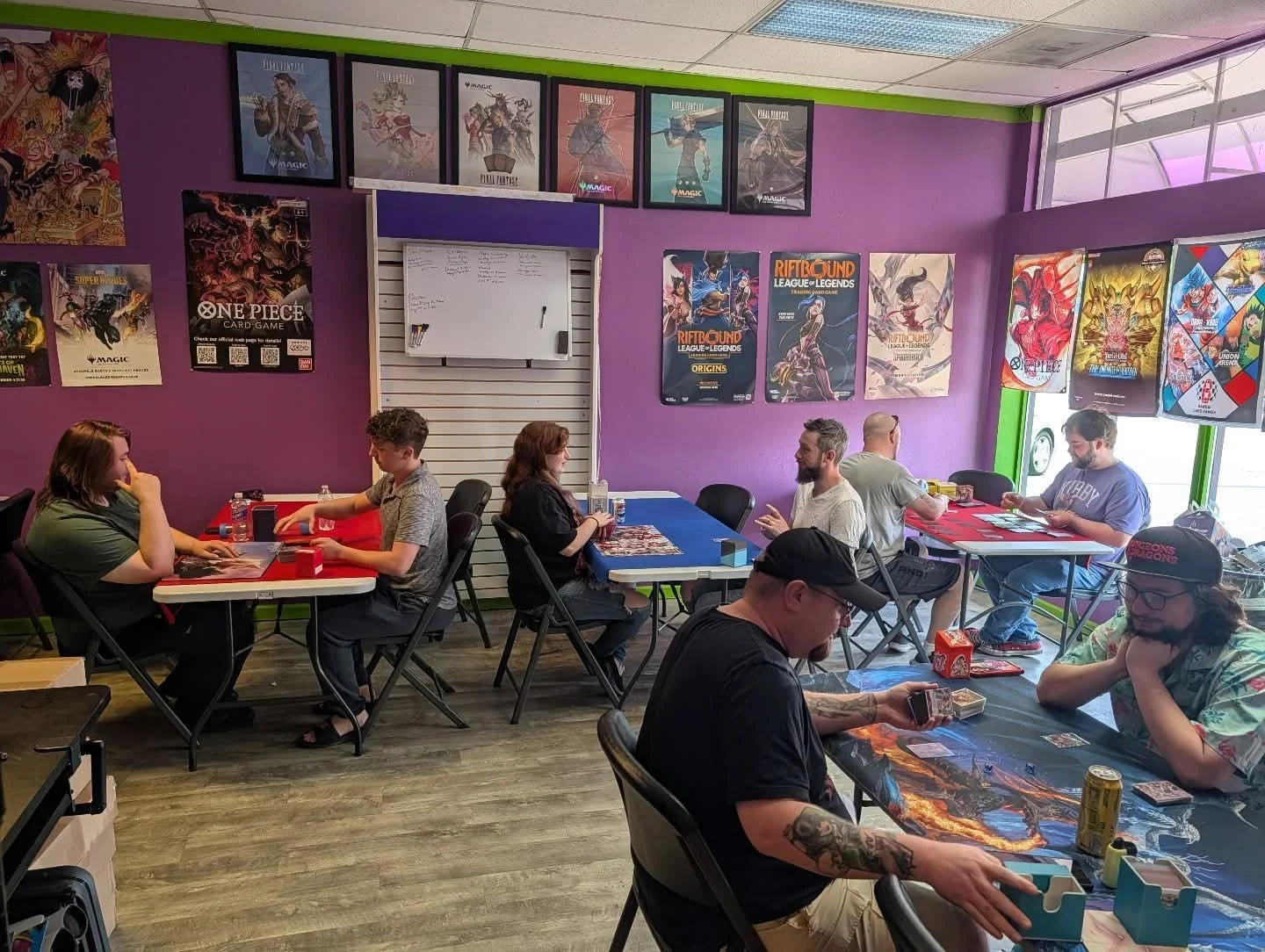 Players gathered at tables for tabletop card games inside a colorful game store decorated with trading card posters.