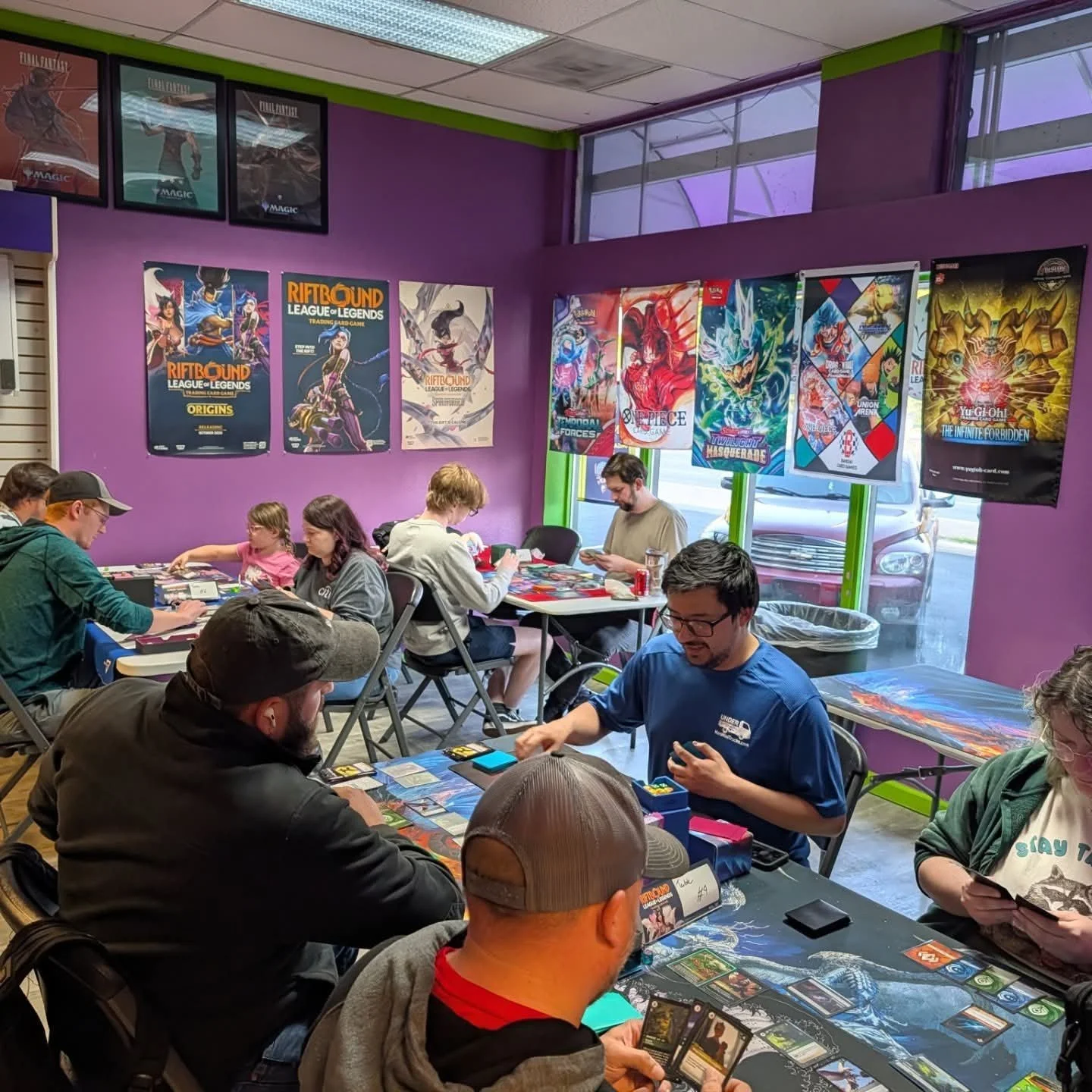 Players gathered around tables for a trading card game event in a colorful game store decorated with Magic: The Gathering, Riftbound, One Piece, and Yu-Gi-Oh! posters.