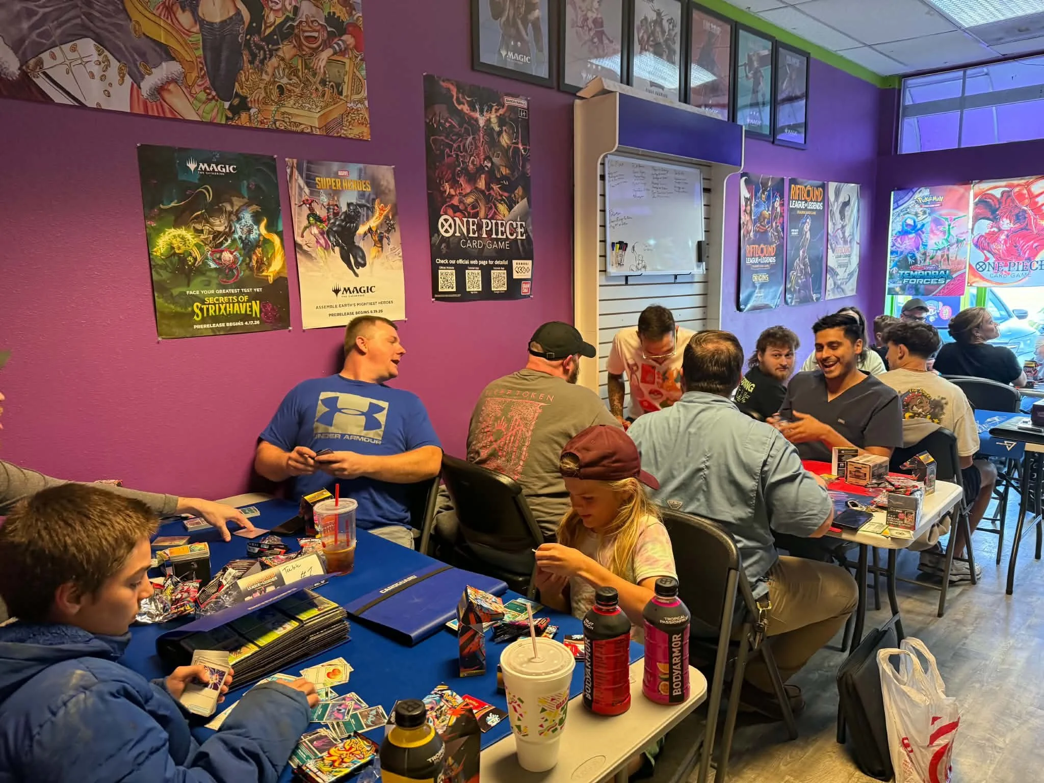 Players gather at tables for trading card games in a colorful game store decorated with Magic: The Gathering, Pokémon, One Piece, and other game posters.