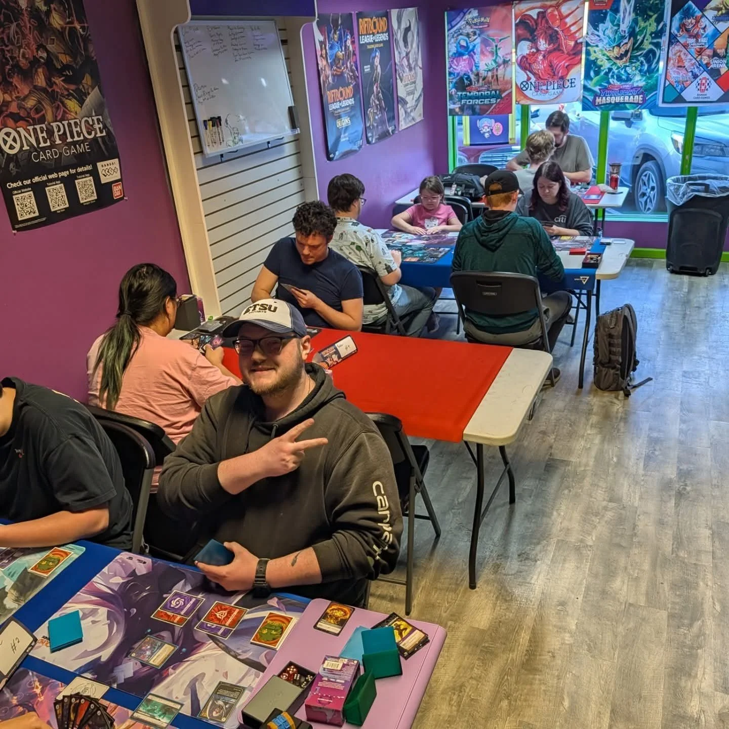 Players gather for trading card games at tables in a colorful game store decorated with One Piece, Pokémon, and League of Legends posters.