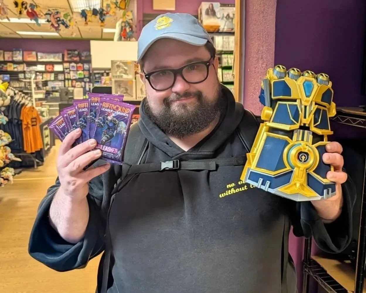 Bearded man in glasses and a blue cap holds League of Legends Riftbound booster packs and a large blue-and-gold fantasy weapon replica in a gaming store.