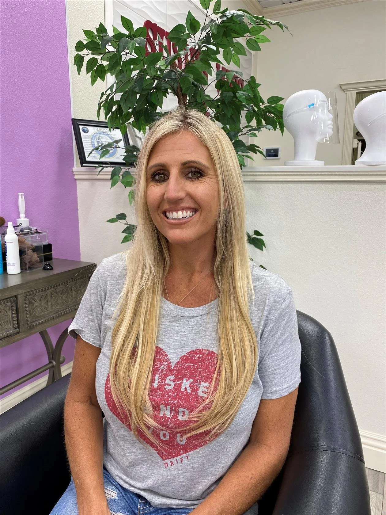 A blonde woman smiling, sitting in a black chair in a salon or beauty studio, wearing a gray T-shirt with a red heart graphic and blue jeans, with framed certificate, plants, and mannequin heads visible in the background.