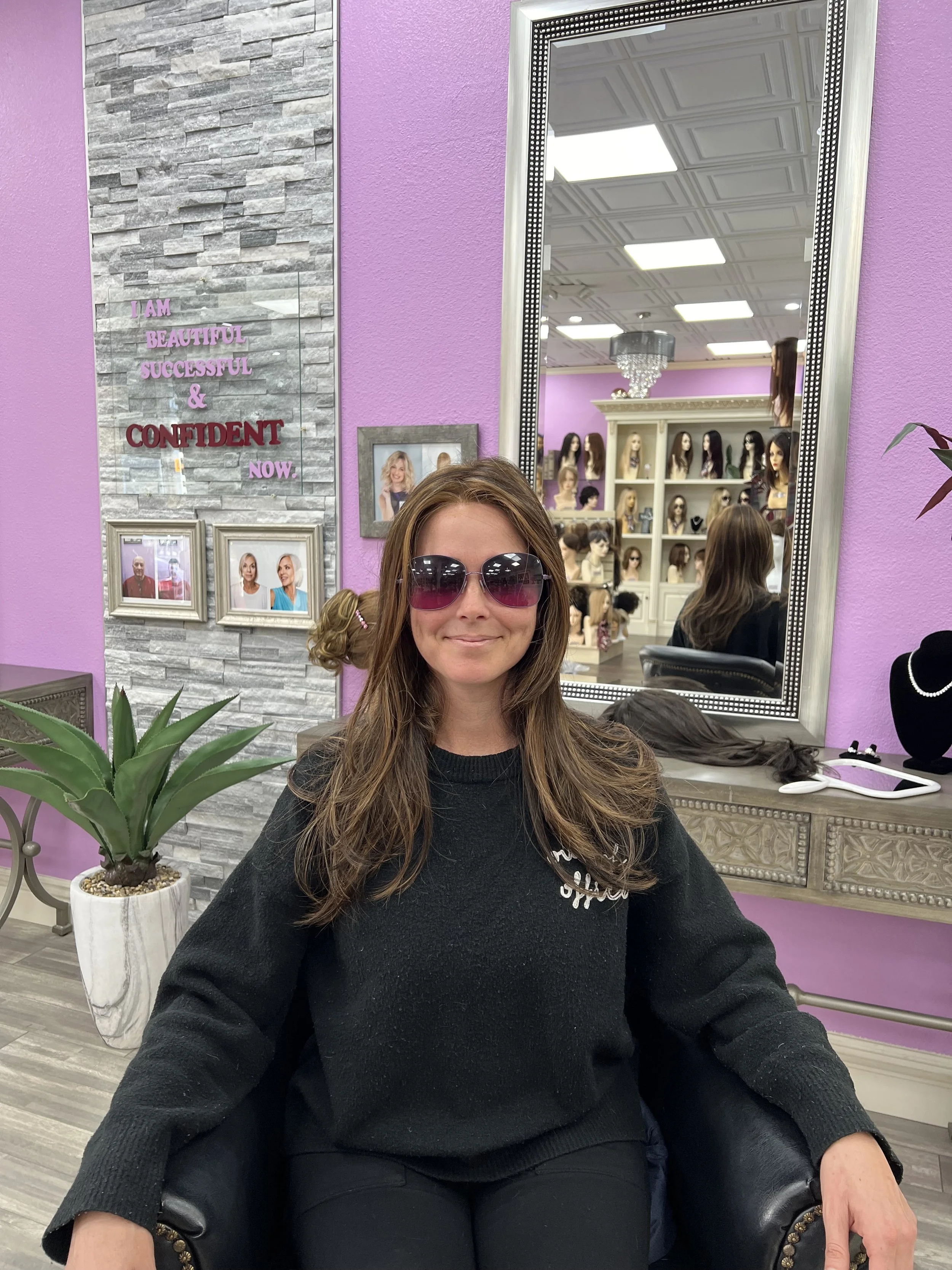 A woman with long brown hair and sunglasses sitting in a salon chair, smiling, in front of a purple wall with a large mirror and shelves of wigs and display mannequins.