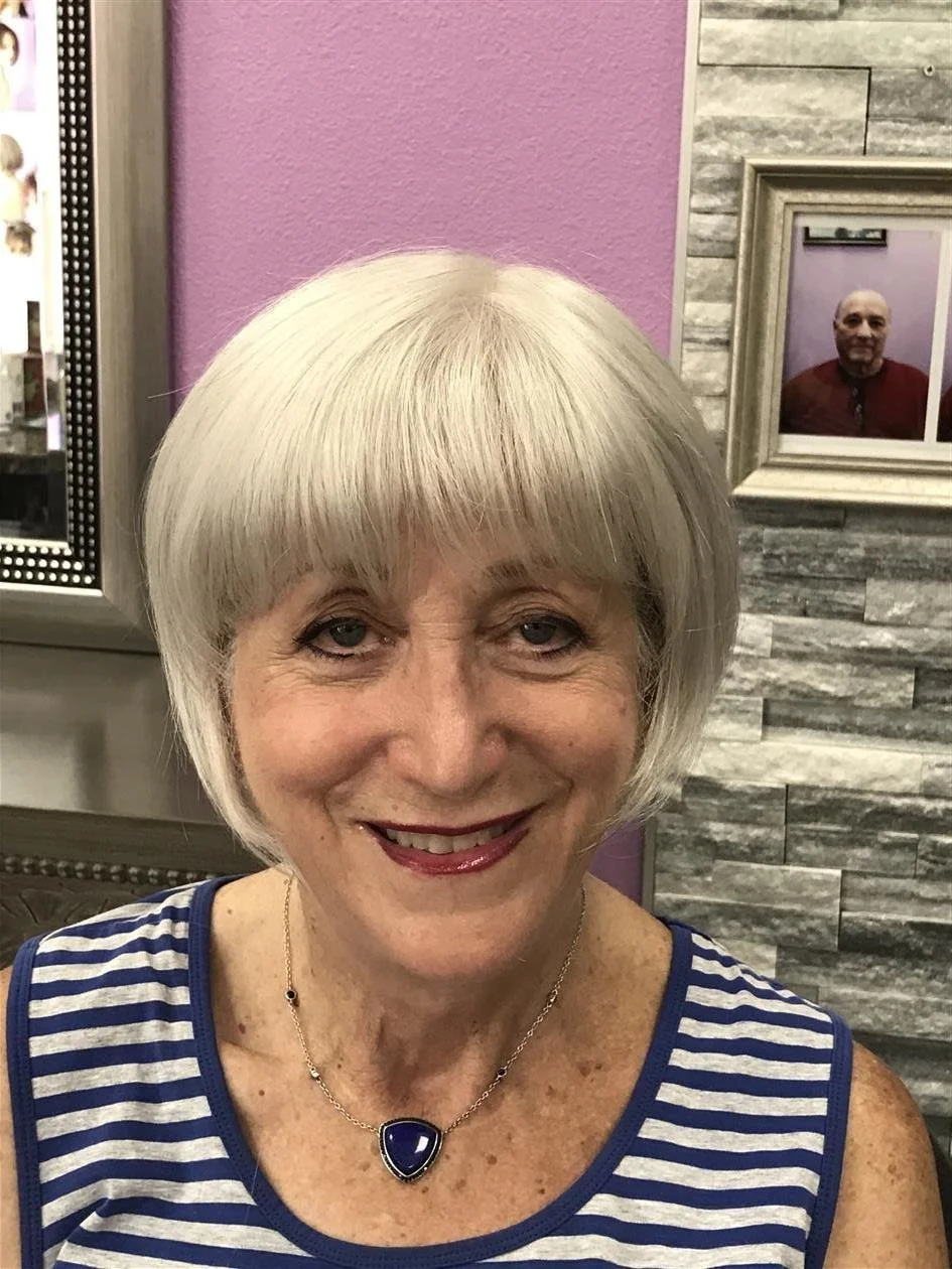 A smiling elderly woman with short white hair, wearing a striped blue and white sleeveless top and a navy heart pendant necklace, sitting in a salon or similar setting.