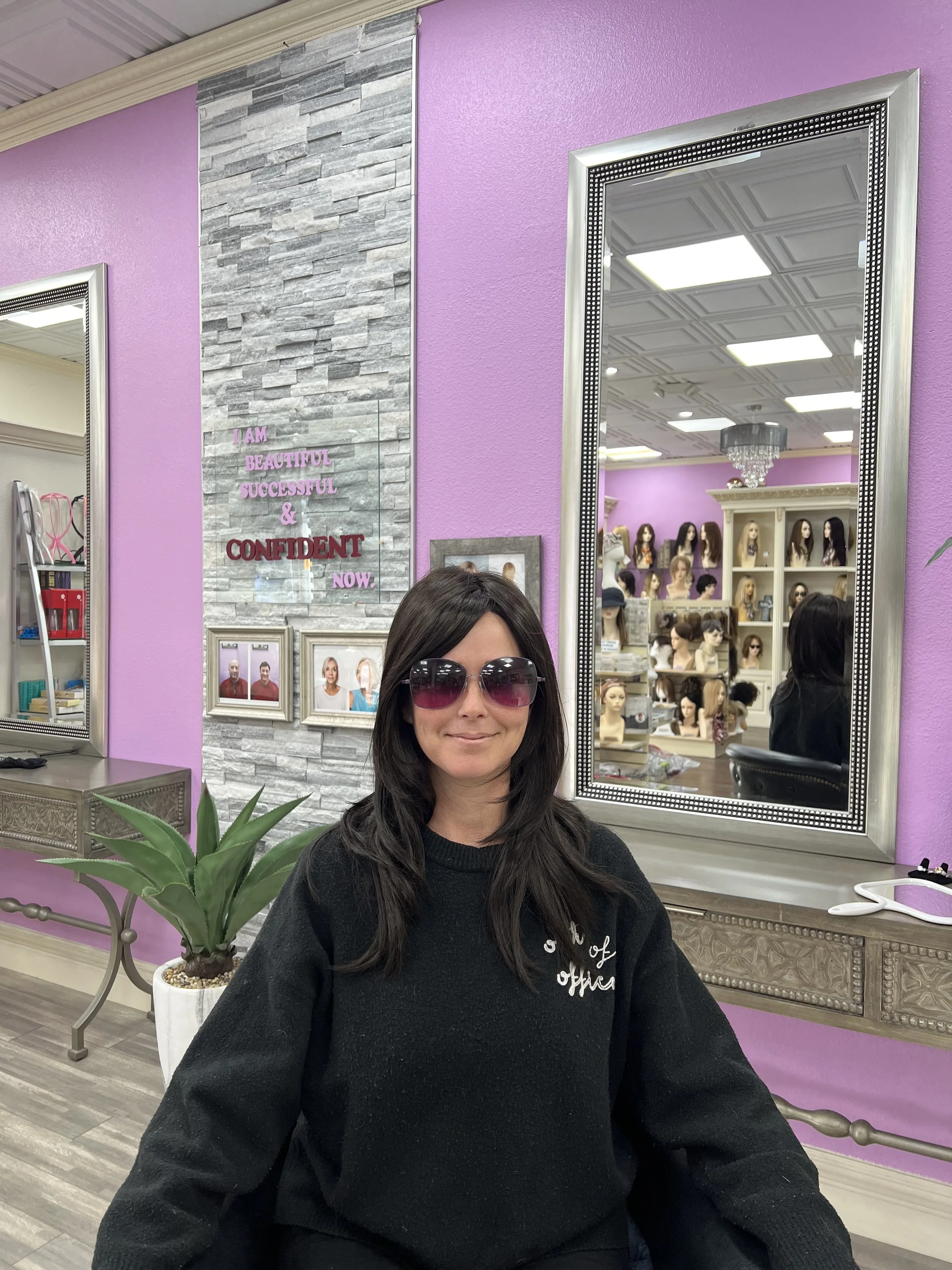 A woman wearing black sweater and sunglasses smiling while sitting in a hair or wig store with purple walls, large mirrors, and display mannequins with various wigs in the background.