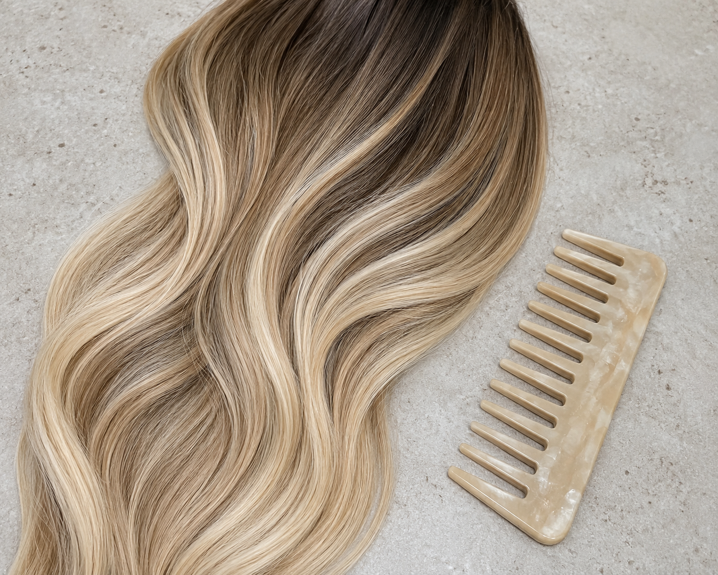 Blonde hair extension with loose waves and a beige hair comb on a light gray surface.