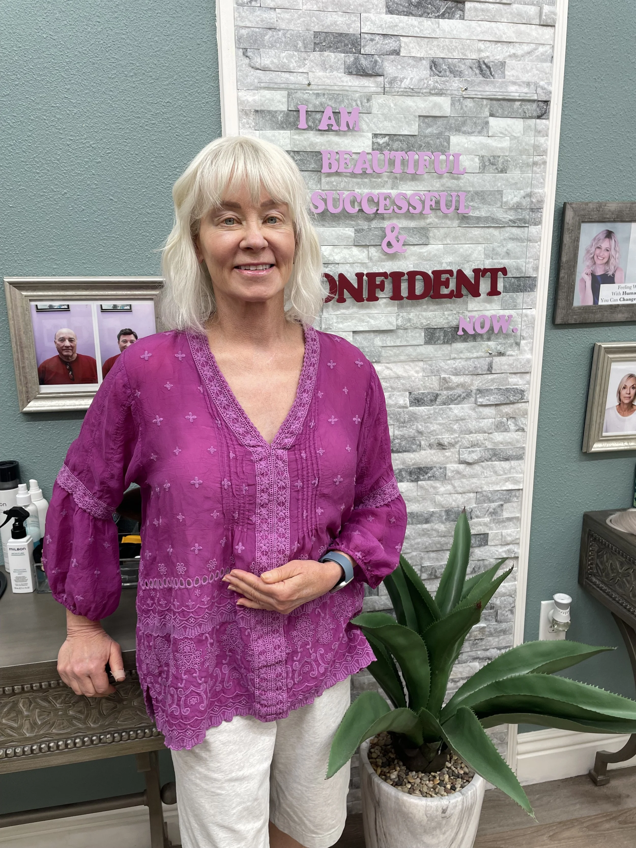 A woman with blonde hair and a pink embroidered blouse standing indoors next to a potted agave plant, with framed photos and a motivational sign on the wall behind her.