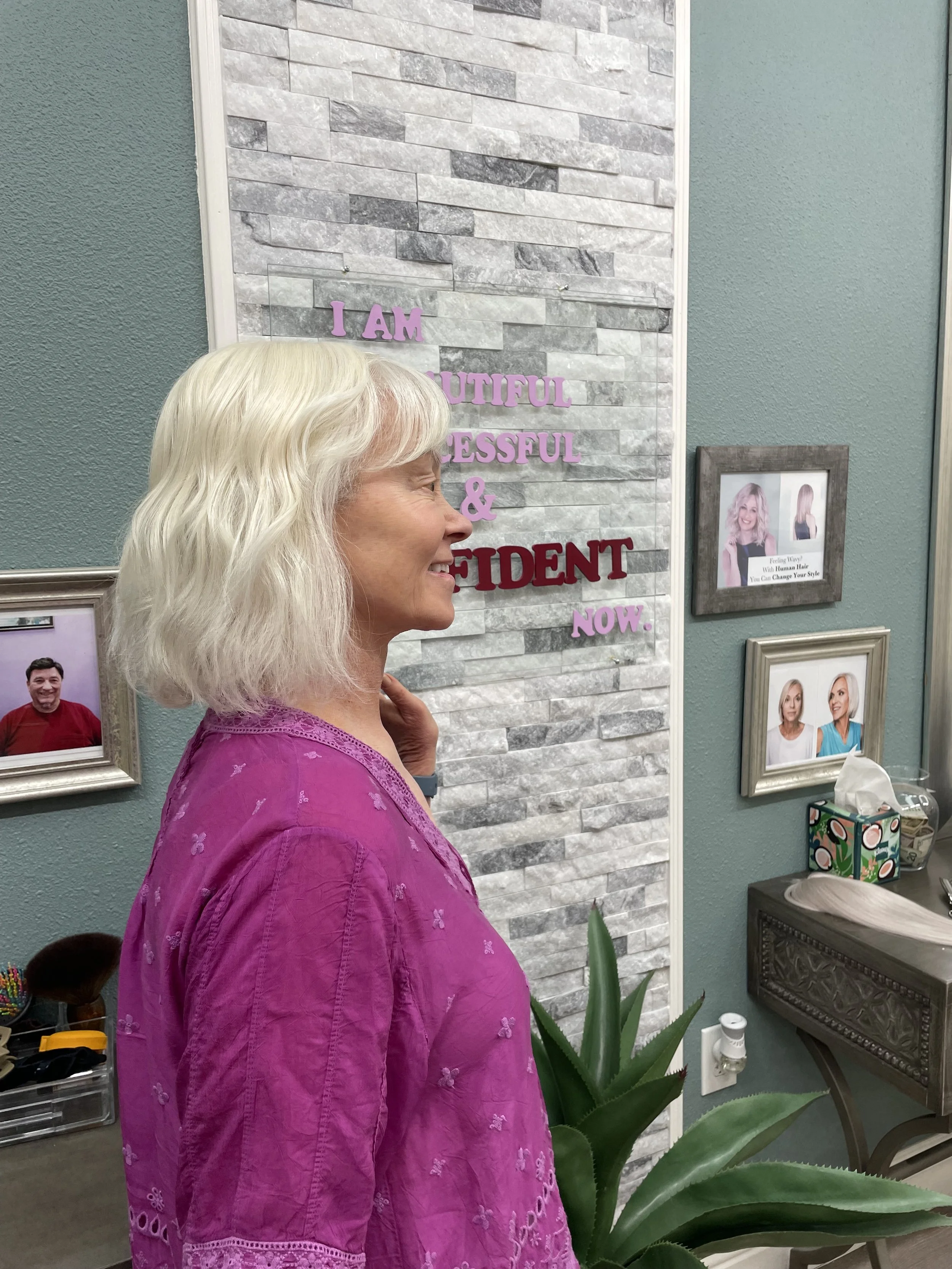 Side profile of a smiling woman with white blonde hair wearing a pink embroidered blouse, standing indoors with framed photographs and decor on the wall behind her, a green plant in front of her.