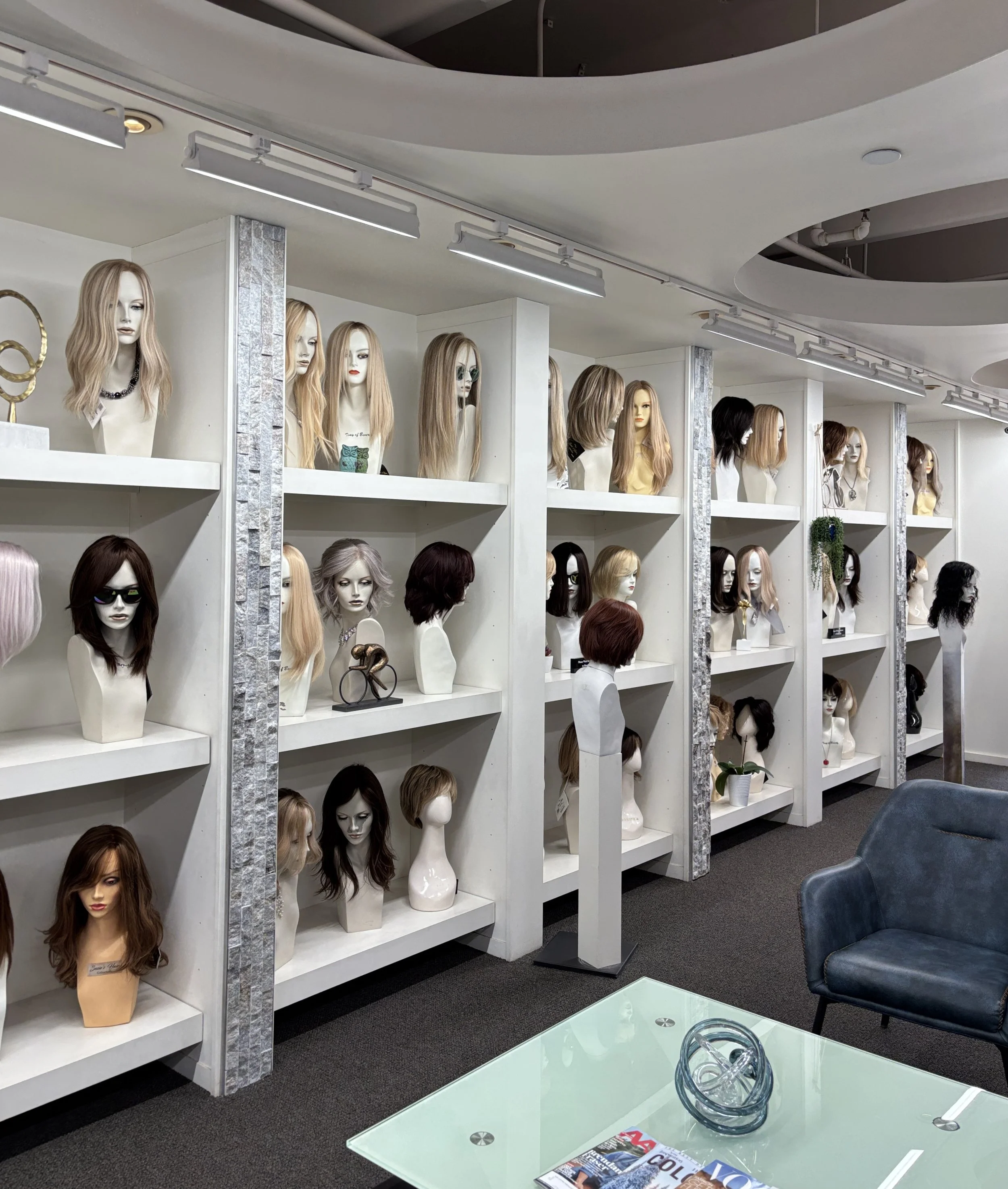 Display of mannequin heads with various long, medium, and short hairstyles, some with wigs and accessories, in a hair salon or wig shop. Shelves and mannequins showcase different hair colors and styles.