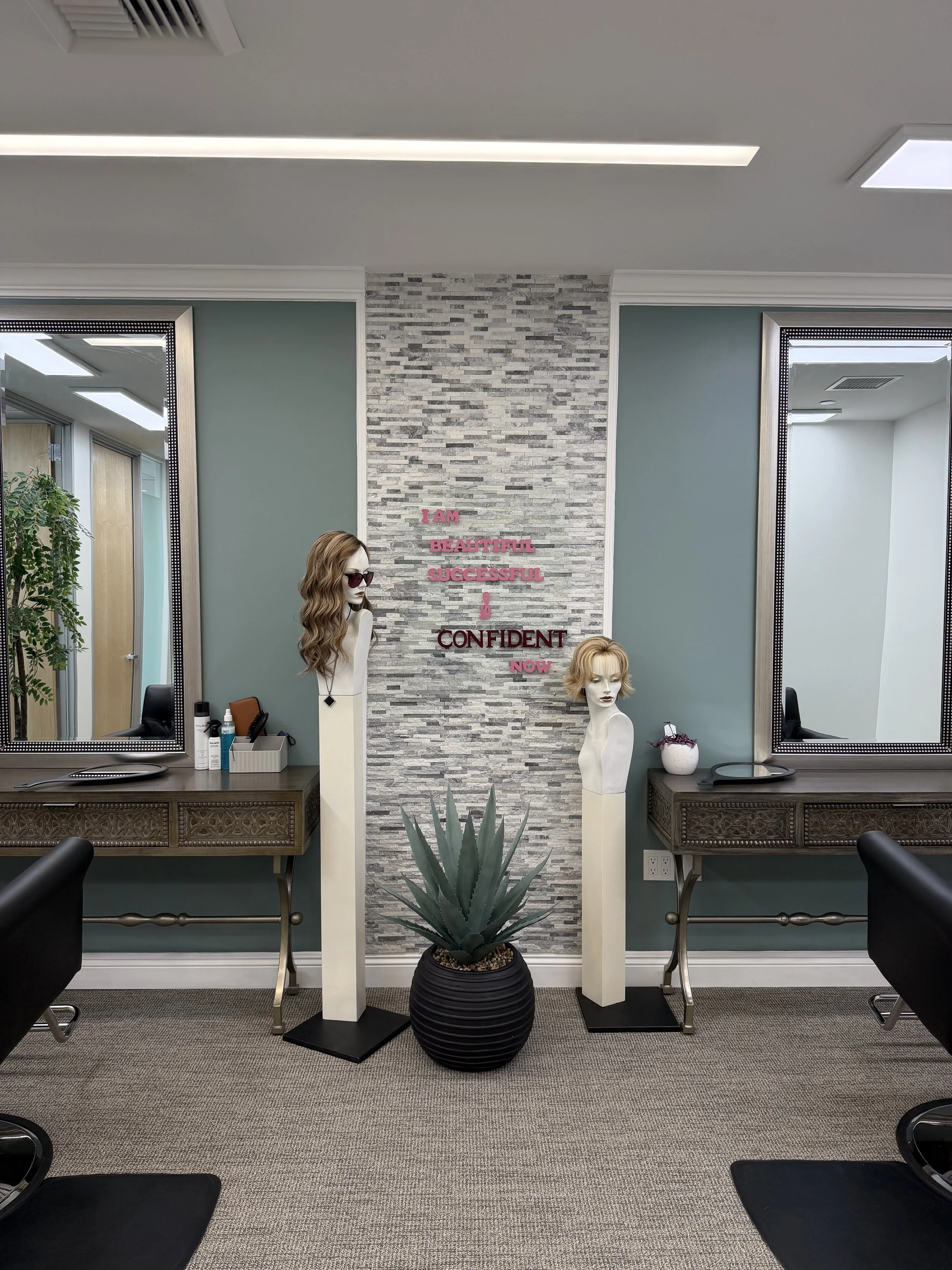 Salon with two mirrors above two desks, a potted plant, and mannequins with wigs, behind a stone accent wall with pink and maroon inspirational words.