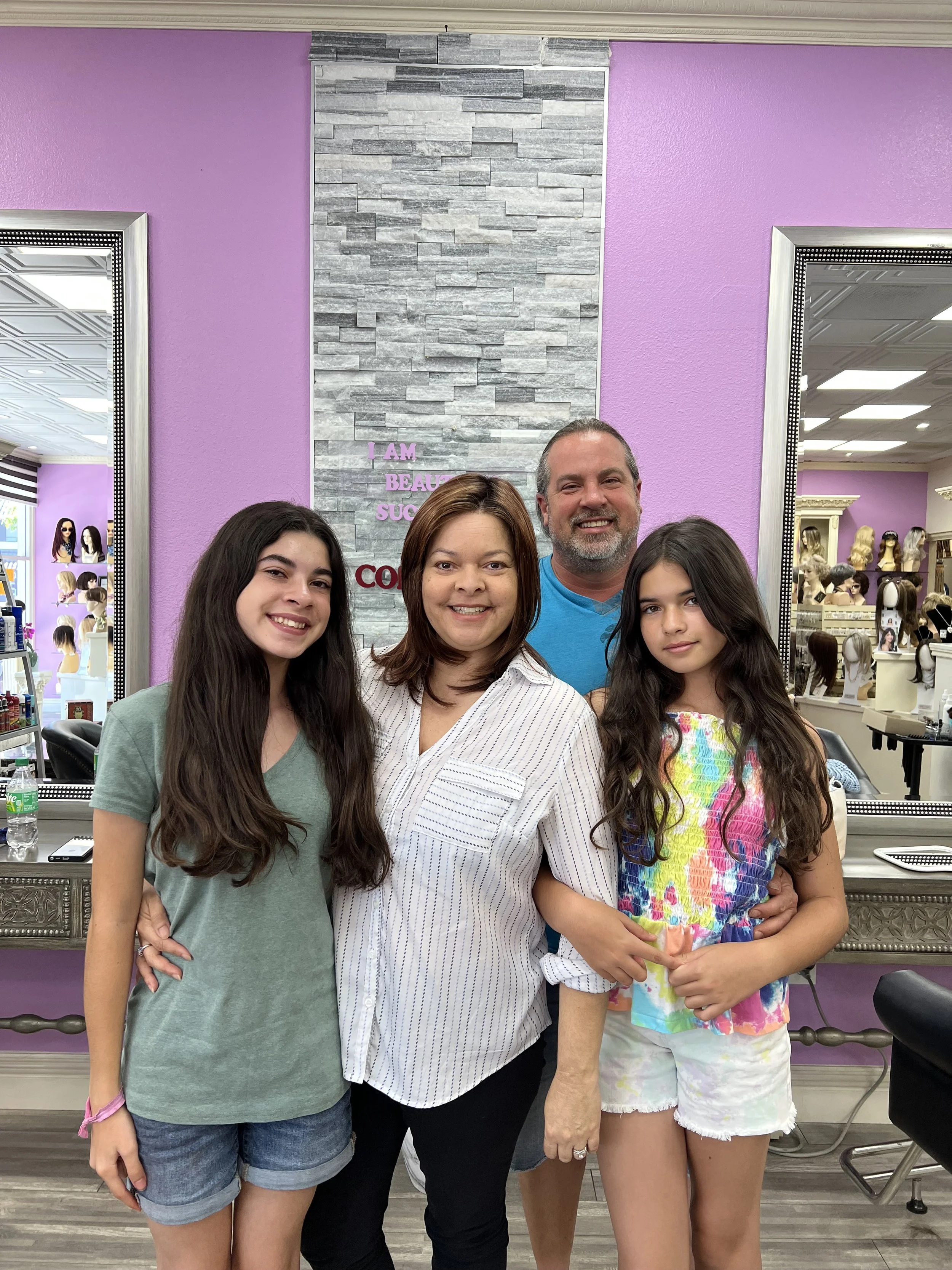 A group of four people standing in a beauty store, smiling at the camera, with purple walls and display mirrors behind them. Two young girls, an older woman, and an older man.