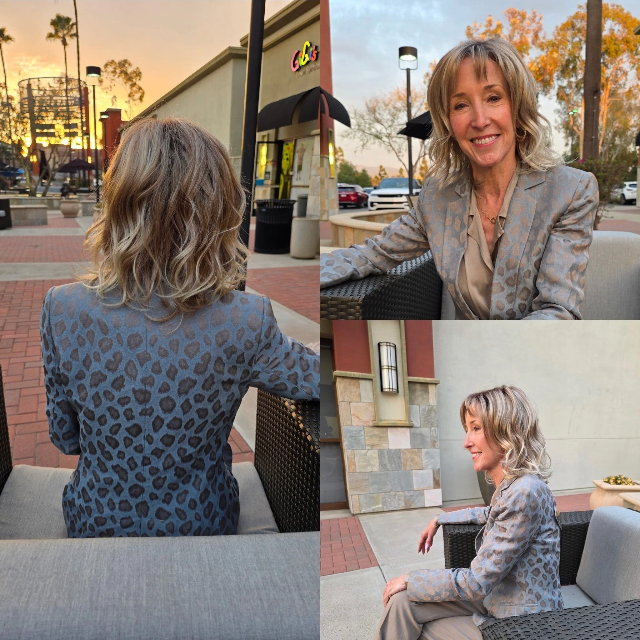 Woman with curly blonde hair sitting outside at a table, smiling, with a building and a parking lot in the background during sunset.