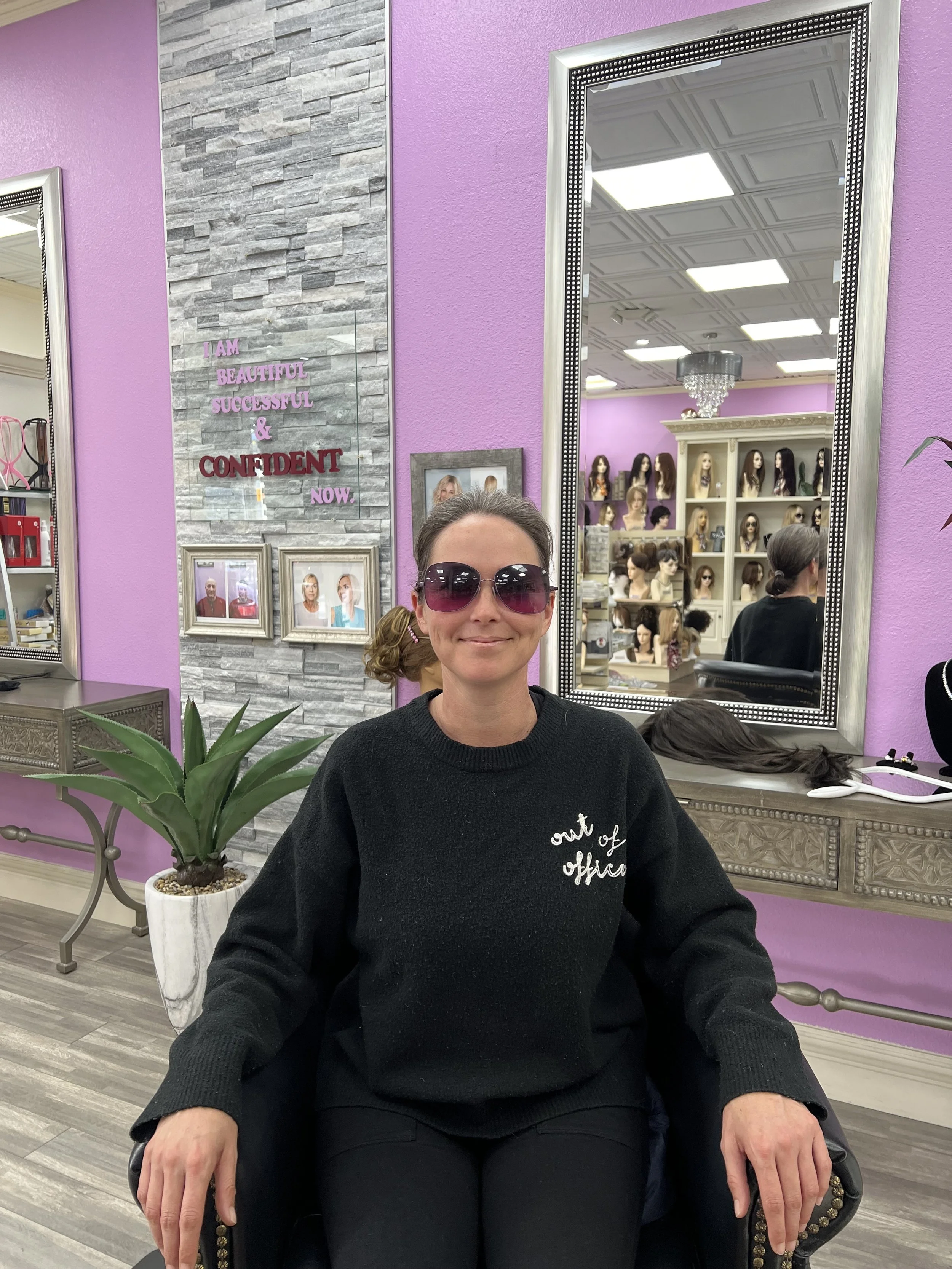 A woman wearing sunglasses, sitting in a salon with pink walls, a large mirror, and a display of mannequin heads with hair in the background.