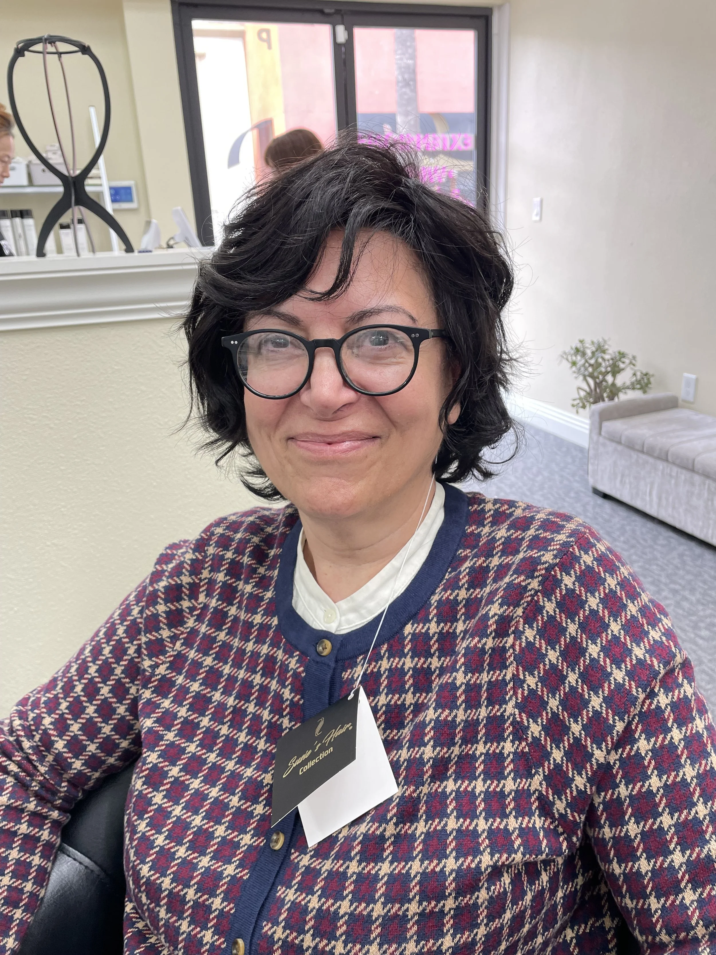 A woman with short wavy black hair and glasses sitting in a salon or office space, wearing a red and blue patterned cardigan and a white collared shirt, smiling at the camera.
