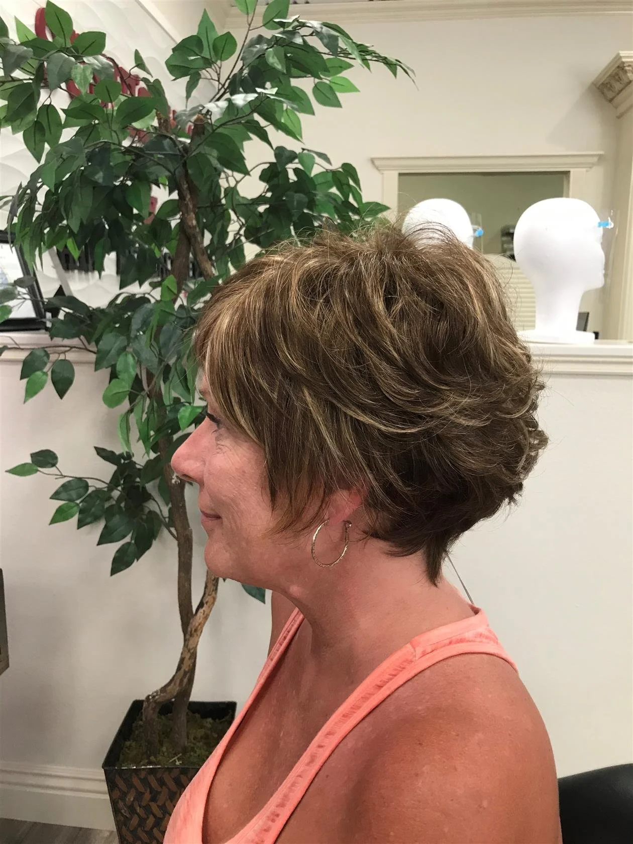 A woman with short, layered brown hair with highlights, wearing earrings and a peach tank top, sitting indoors with a large green plant and white mannequin heads in the background.