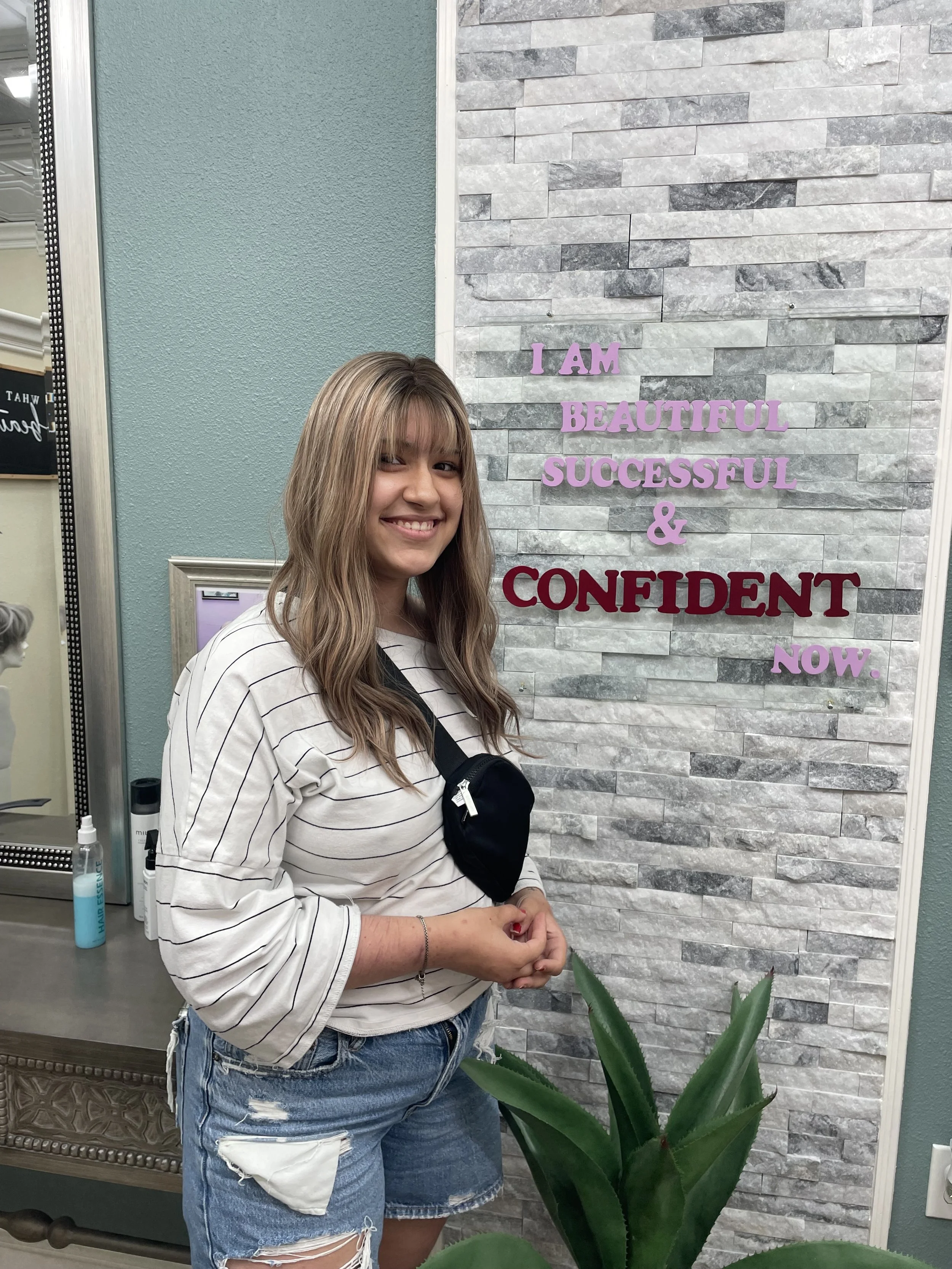 A woman with long light brown hair, wearing a white and black striped long-sleeve shirt and ripped denim shorts, smiling and standing in front of a wall with motivational words in pink and red letters that say 'I am beautiful, successful & confident now.'