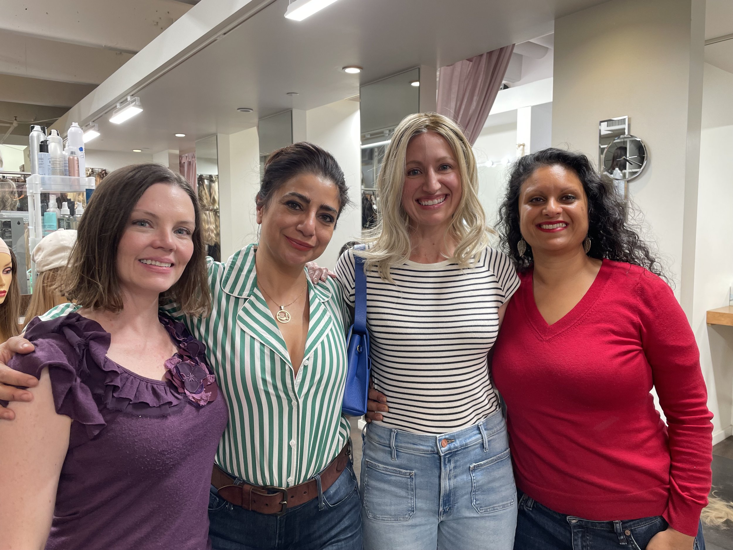 Four women smiling and standing close together indoors, with various hair and clothing styles, and a mirror and display of hair and beauty products in the background.