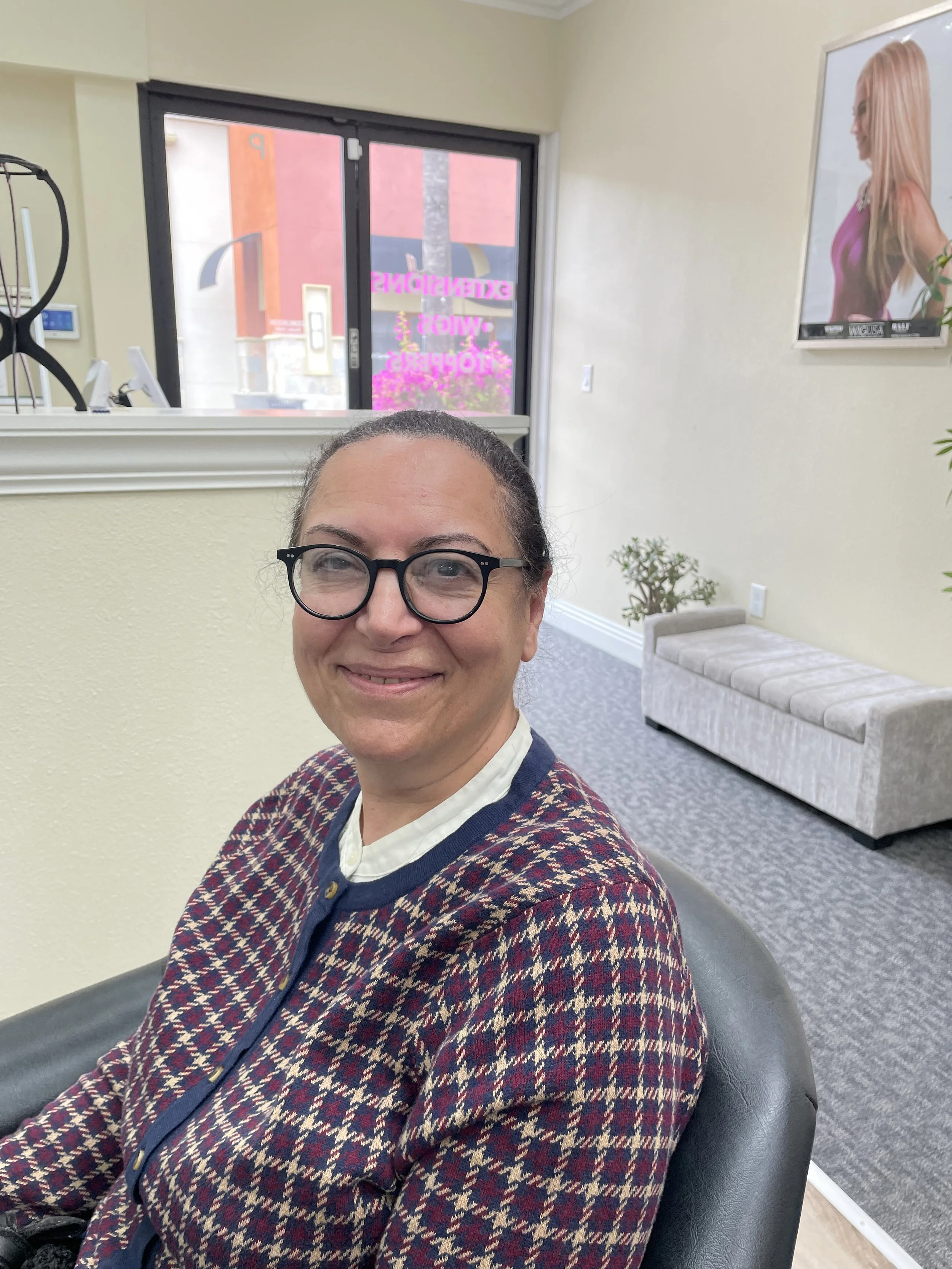 Woman with glasses smiling, sitting in a waiting area inside a professional office. She is wearing a patterned sweater and has brown hair.