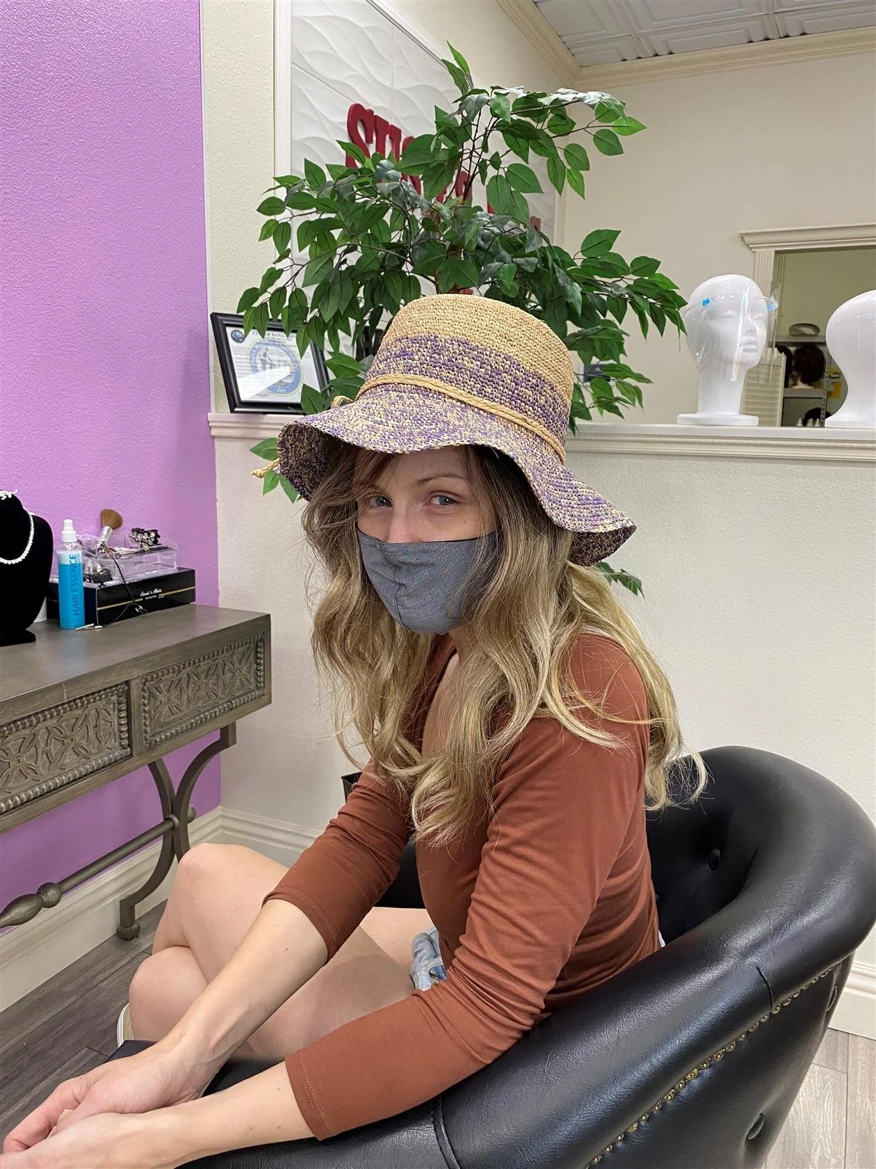 A woman with long wavy blonde hair wearing a large straw sun hat and a gray face mask, sitting in a black chair in a salon or spa. She is dressed in a brown long-sleeve top with her arms resting on her legs. In the background, there is a purple wall with a glass display case, a plant, and decorative mannequins on a shelf.