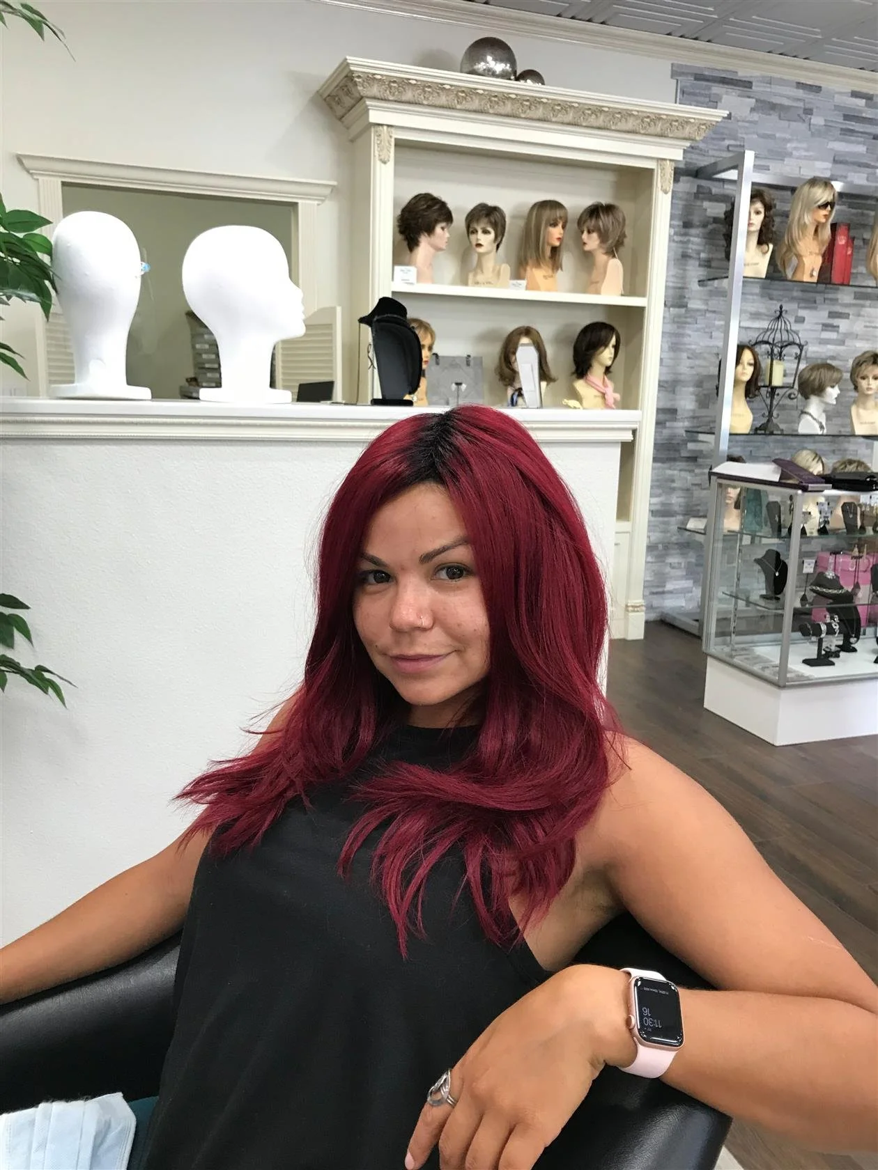 A woman with long, wavy, burgundy-colored hair sitting in a hair salon chair, wearing a black sleeveless top and an Apple Watch on her left wrist. The salon has mannequin heads with various hairstyles on shelves behind her and a mirror reflecting other mannequin heads.