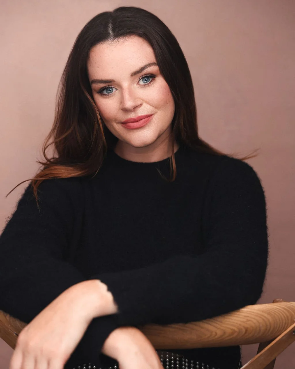 A woman with long dark hair, blue eyes, and fair skin, smiling, wearing a black sweater, sitting at a wooden table with a beige background.