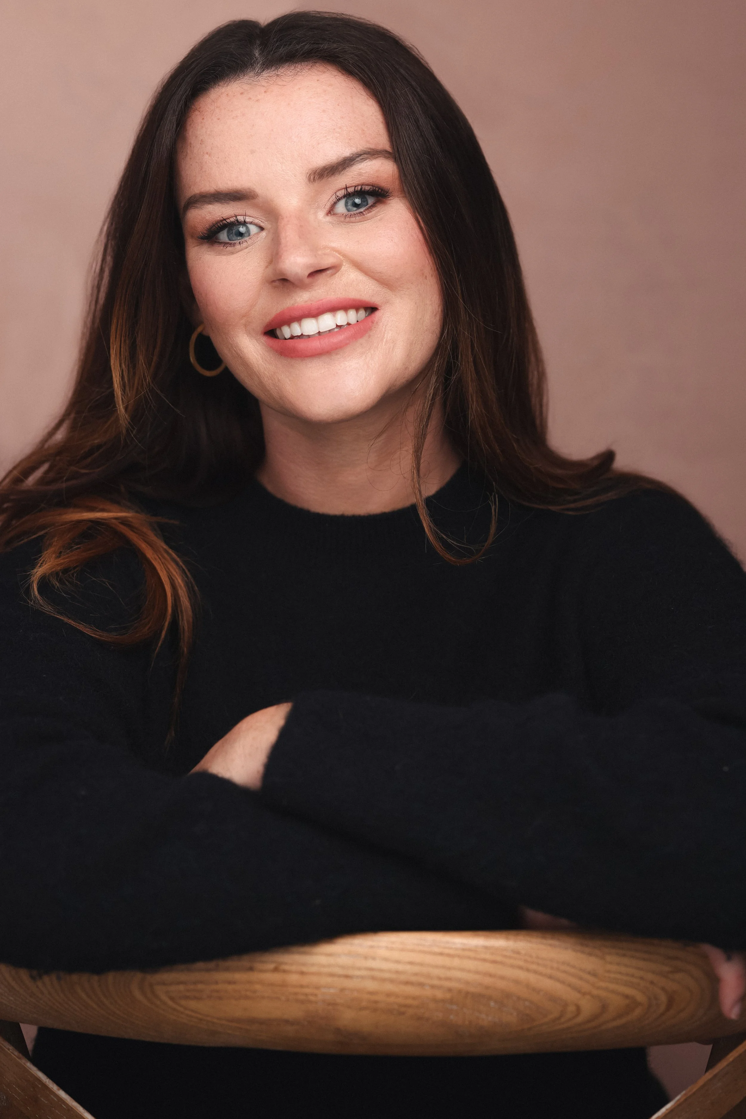 A woman with long brown hair, blue eyes, and wearing a black top, sitting at a wooden table with her arms crossed, smiling at the camera against a plain beige background.