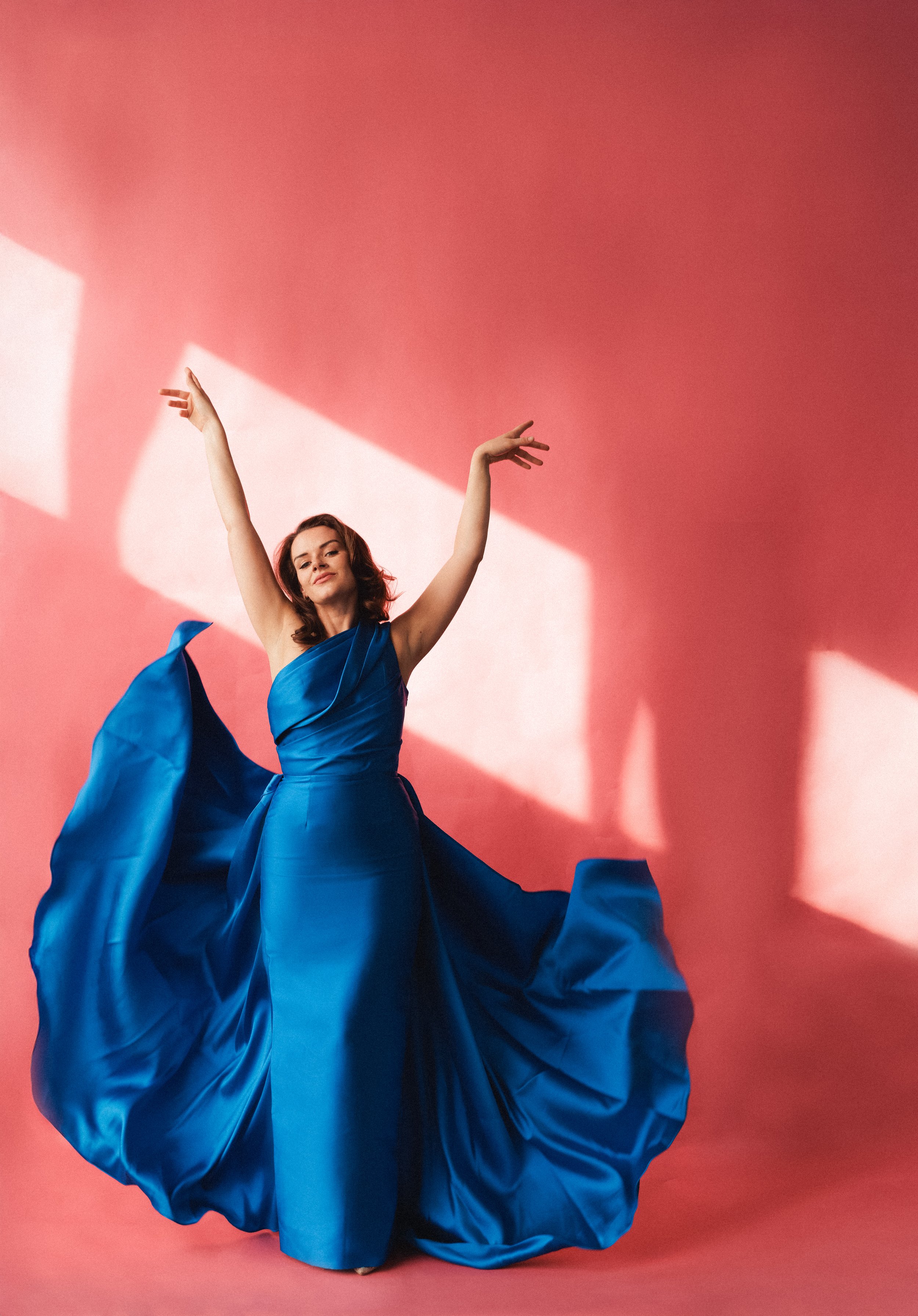 A woman in a flowing blue dress with a ruffled skirt is striking a pose against a pink background with sunlight creating shadows. Her arms are raised and she has a confident expression.