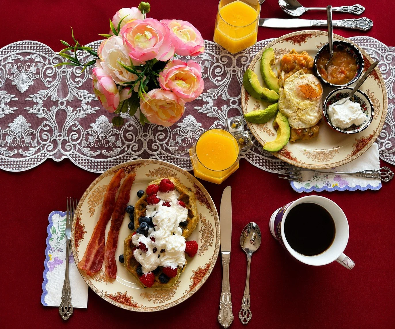A breakfast table with two plates of food, two glasses of orange juice, a cup of coffee, and a pink flower arrangement on a lace tablecloth.