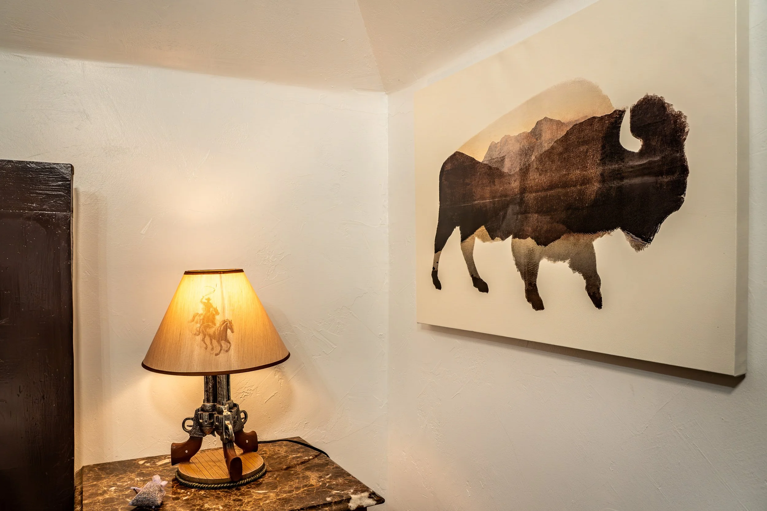 A corner of a room with a lamp on a wooden table, a black gun-shaped base, a small decorative figure, a canvas with a buffalo silhouette and mountain scene, and a wall with textured white paint.