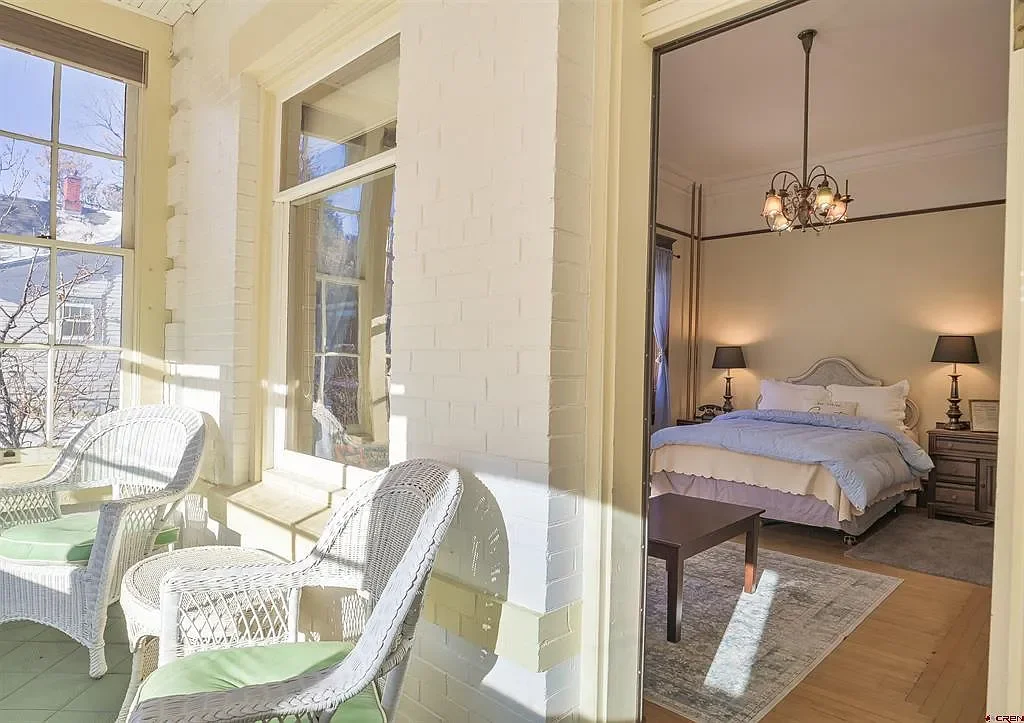 A sunlit porch with white wicker chairs and green cushions, connected to a bedroom with a bed, nightstands, lamps, and a hanging chandelier.